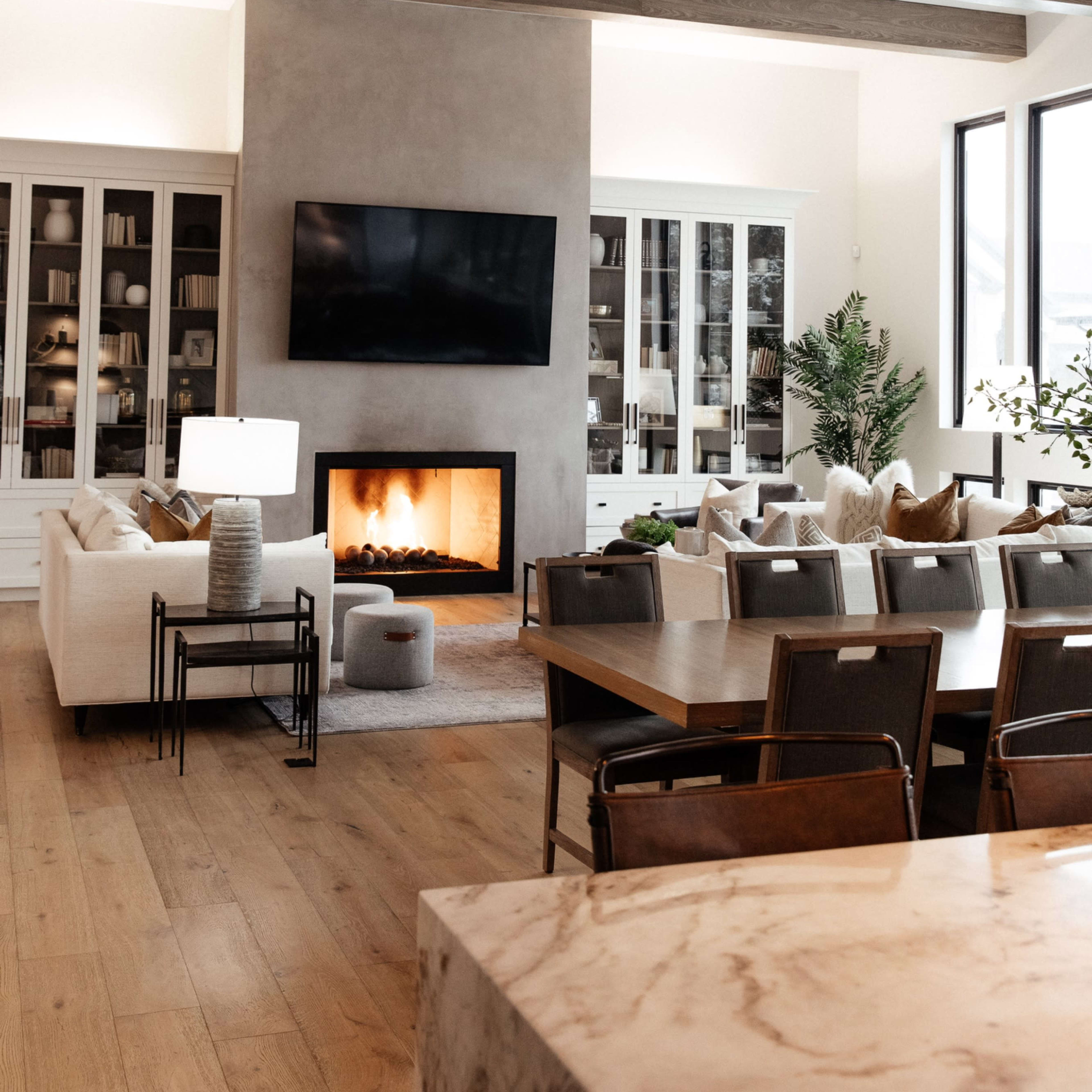 A modern living room featuring a fireplace, large windows, wooden beams on the ceiling, and a mix of comfortable seating arrangements.