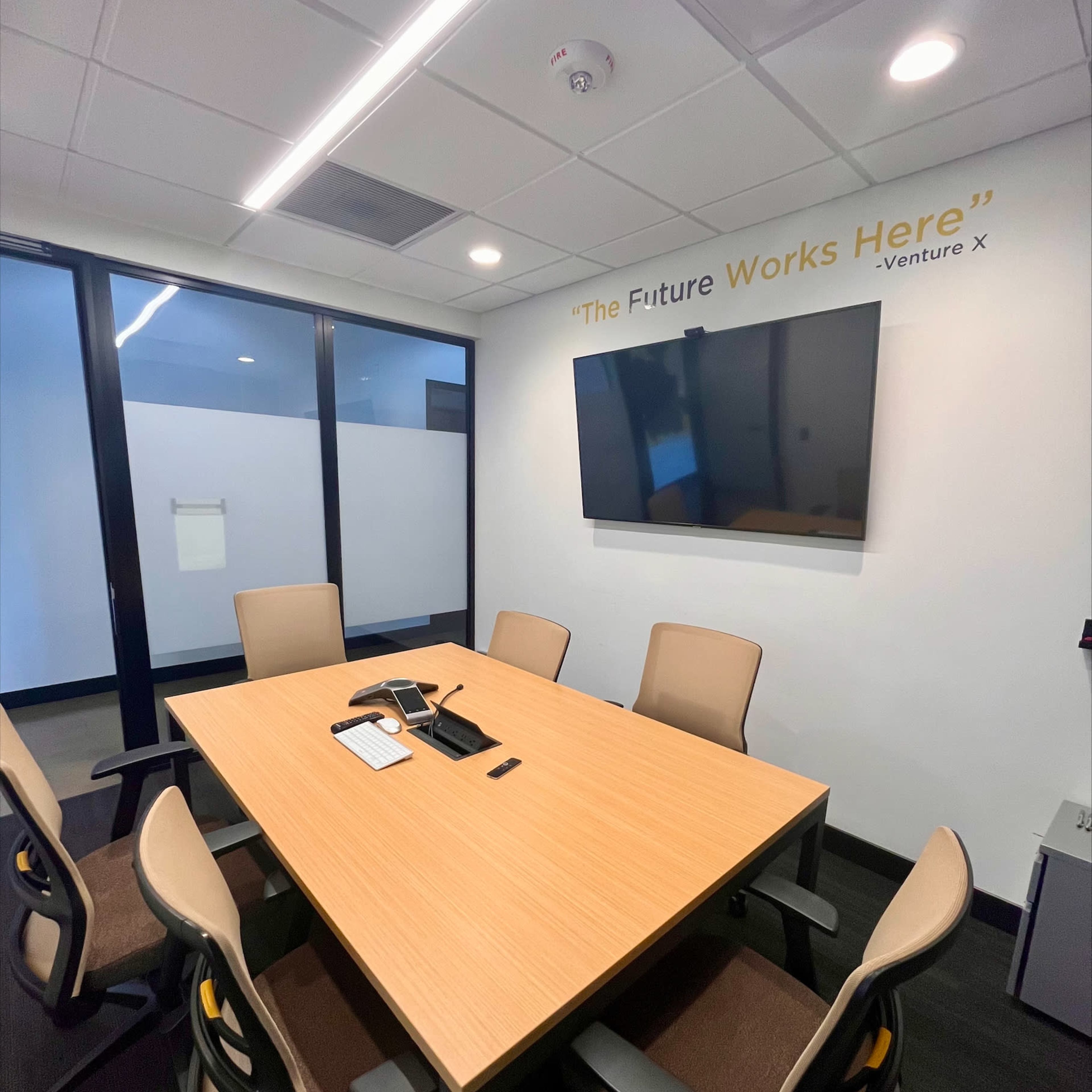 A modern conference room with a wooden table, six chairs, a large TV mounted on the wall, and a motivational quote above the screen.