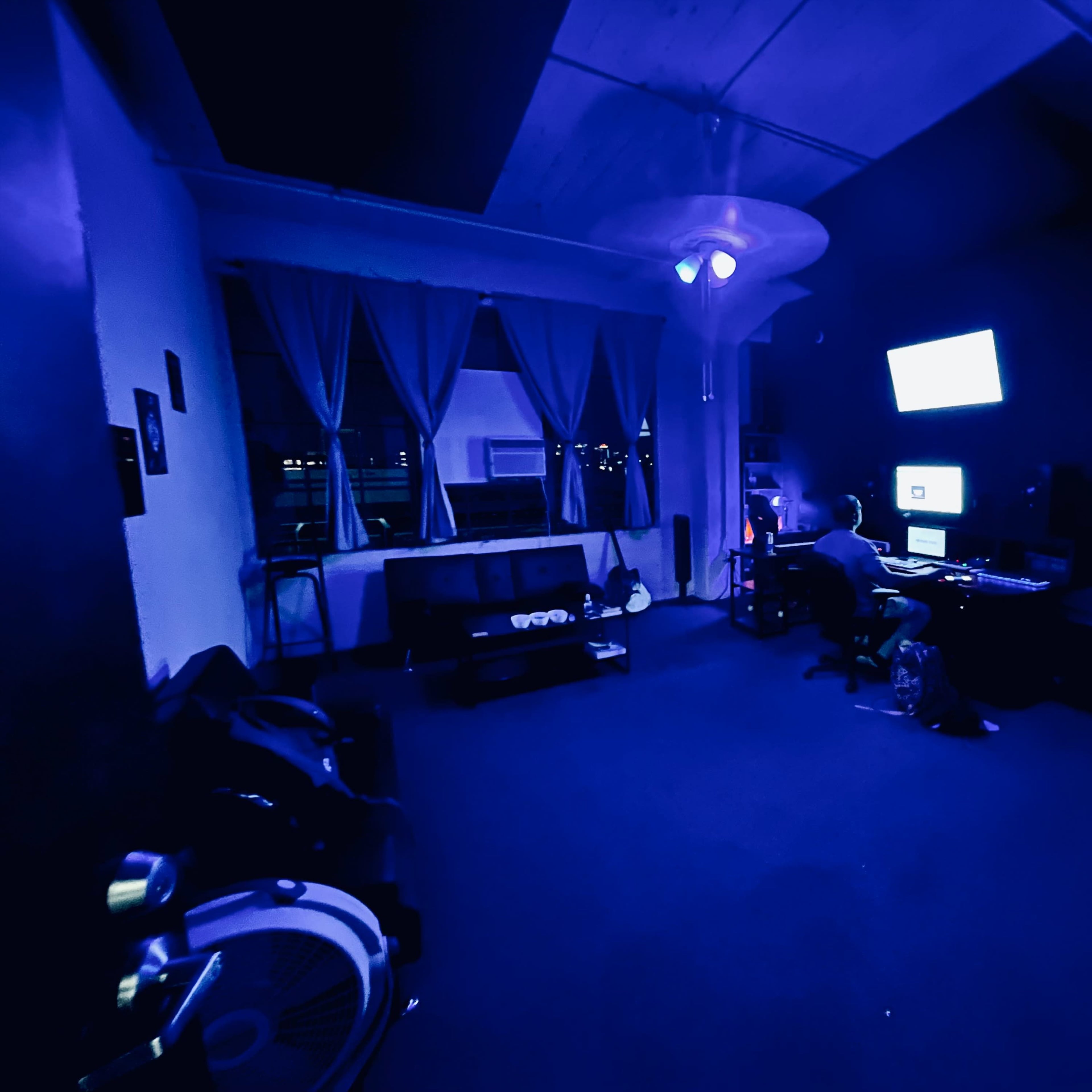 A dimly lit office space with a person sitting at a desk, working on a computer, while a couch and a large window with curtains are present in the background.