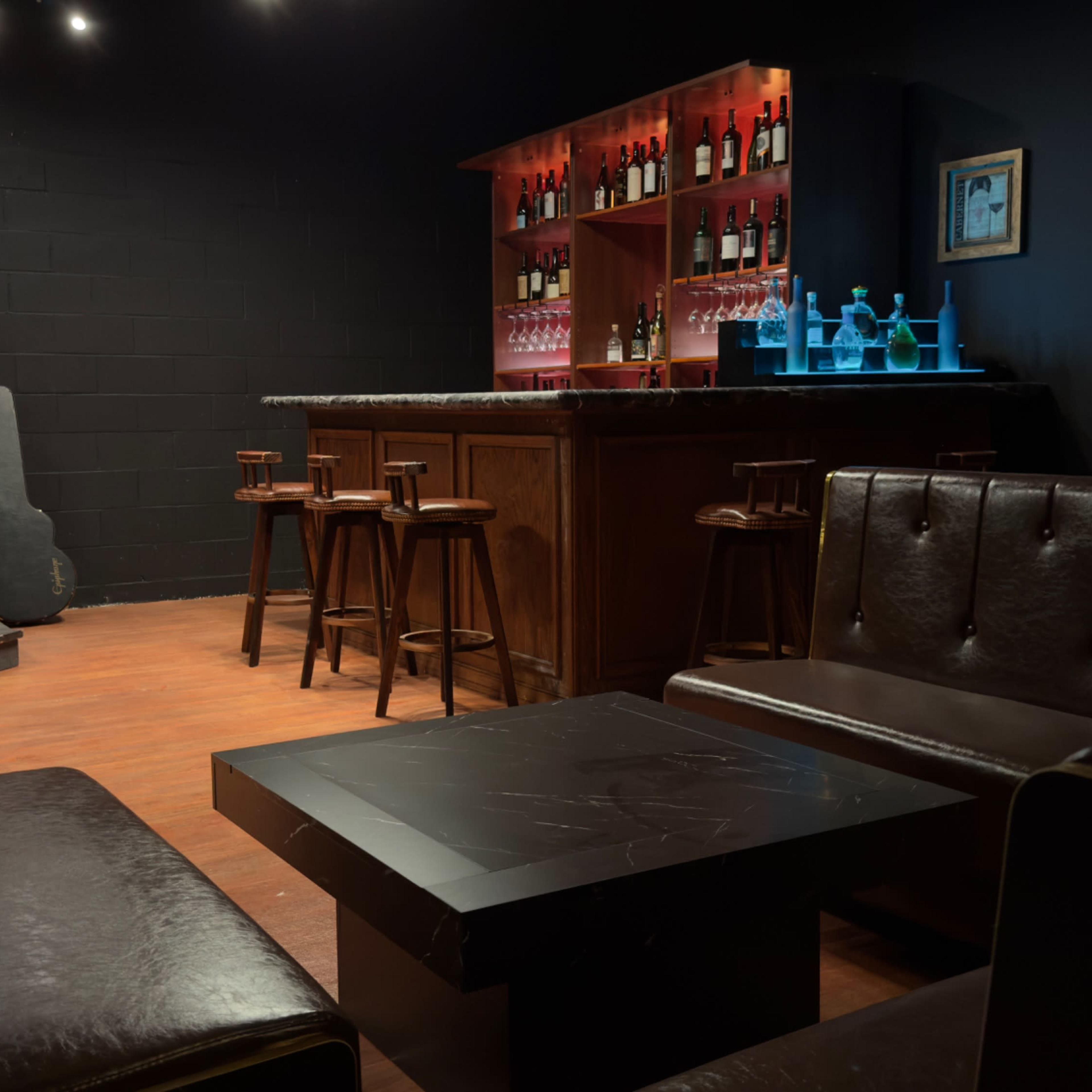 The scene shows a dimly lit bar area with a stage for live music, featuring a wooden bar stocked with drinks, a few high stools, and leather seating arrangements.