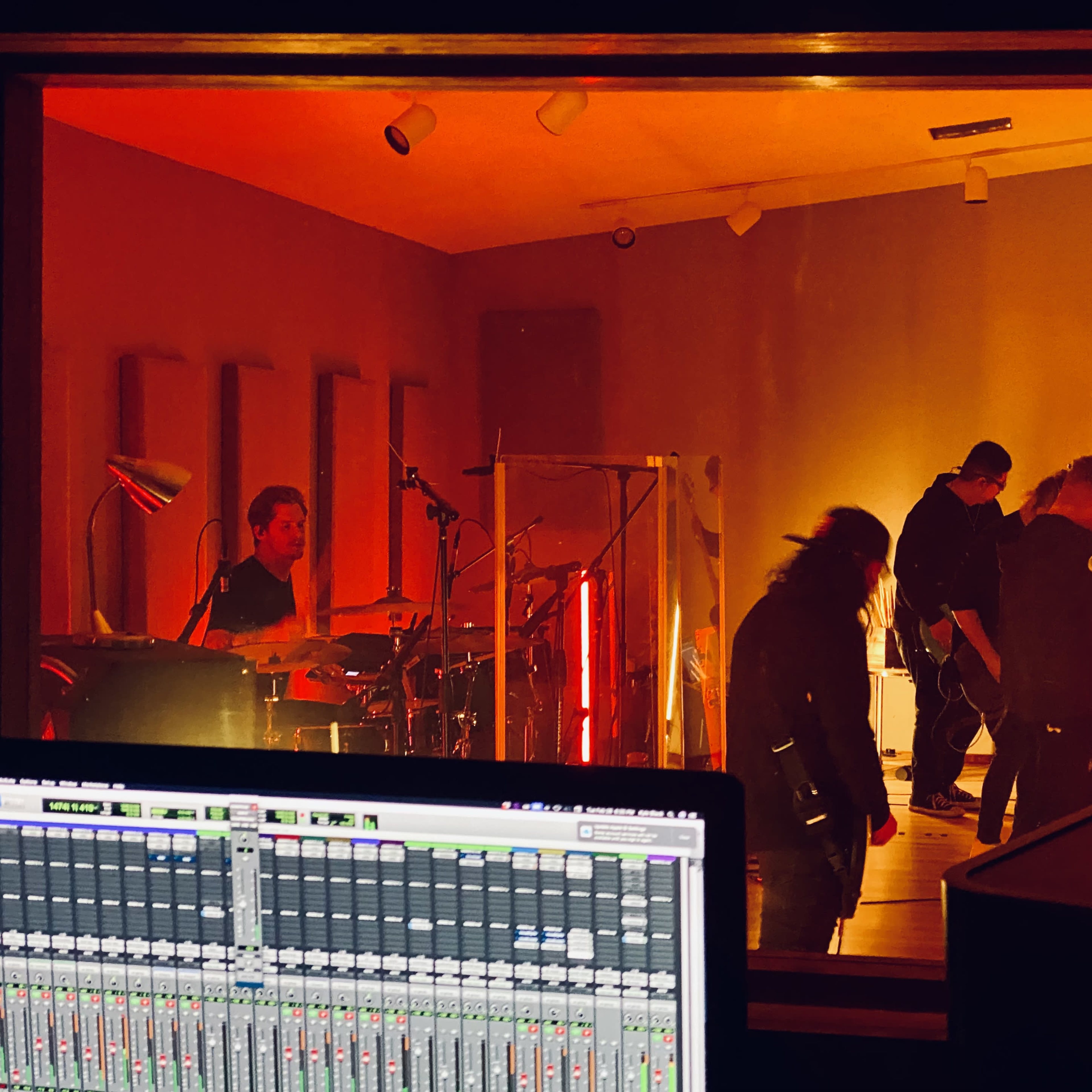 A band performs in a recording studio, illuminated by orange lights, while a mixing console with audio tracks is visible in the foreground.