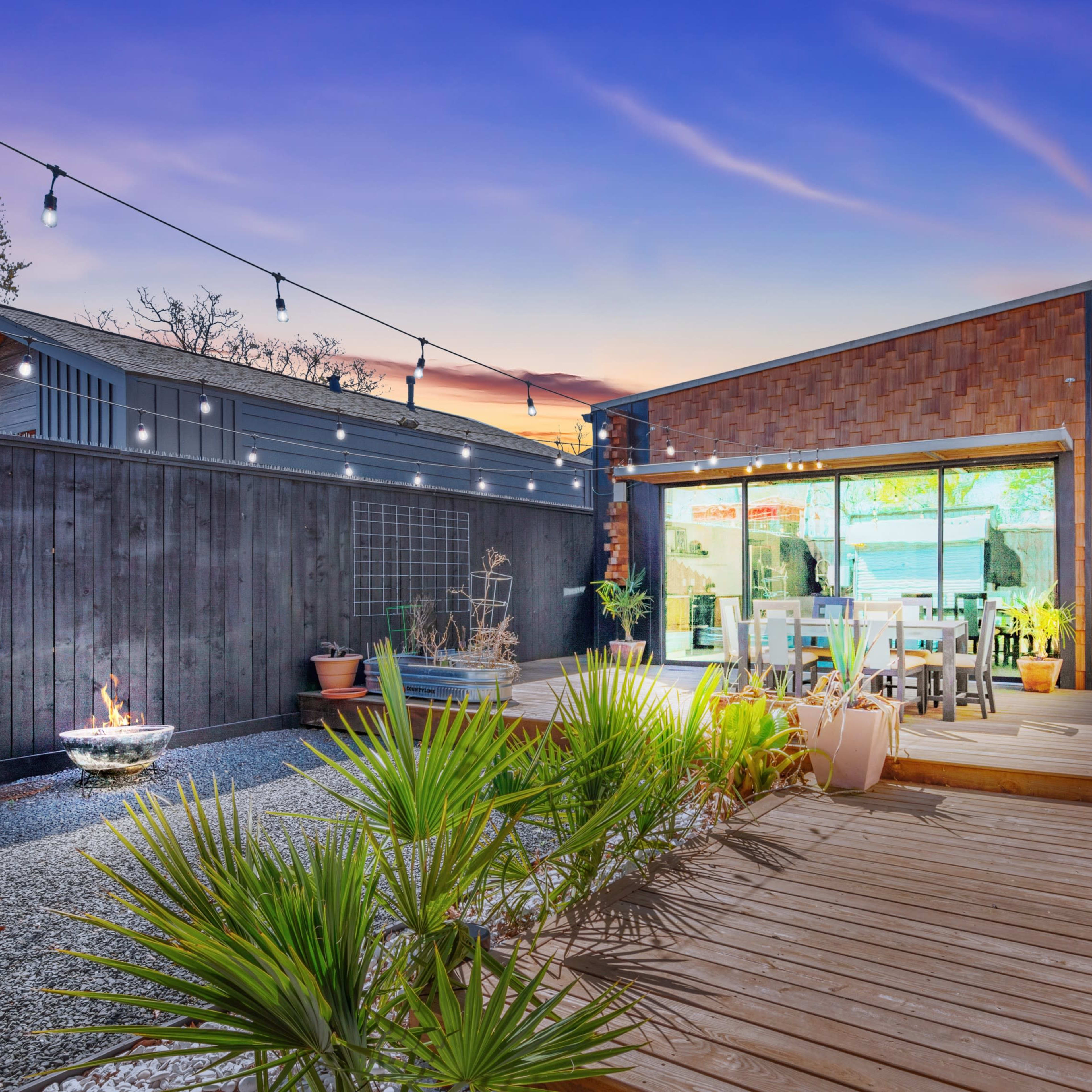 A modern outdoor patio area featuring wooden decking, potted plants, and a small fire pit, with a large glass door leading into a contemporary house.