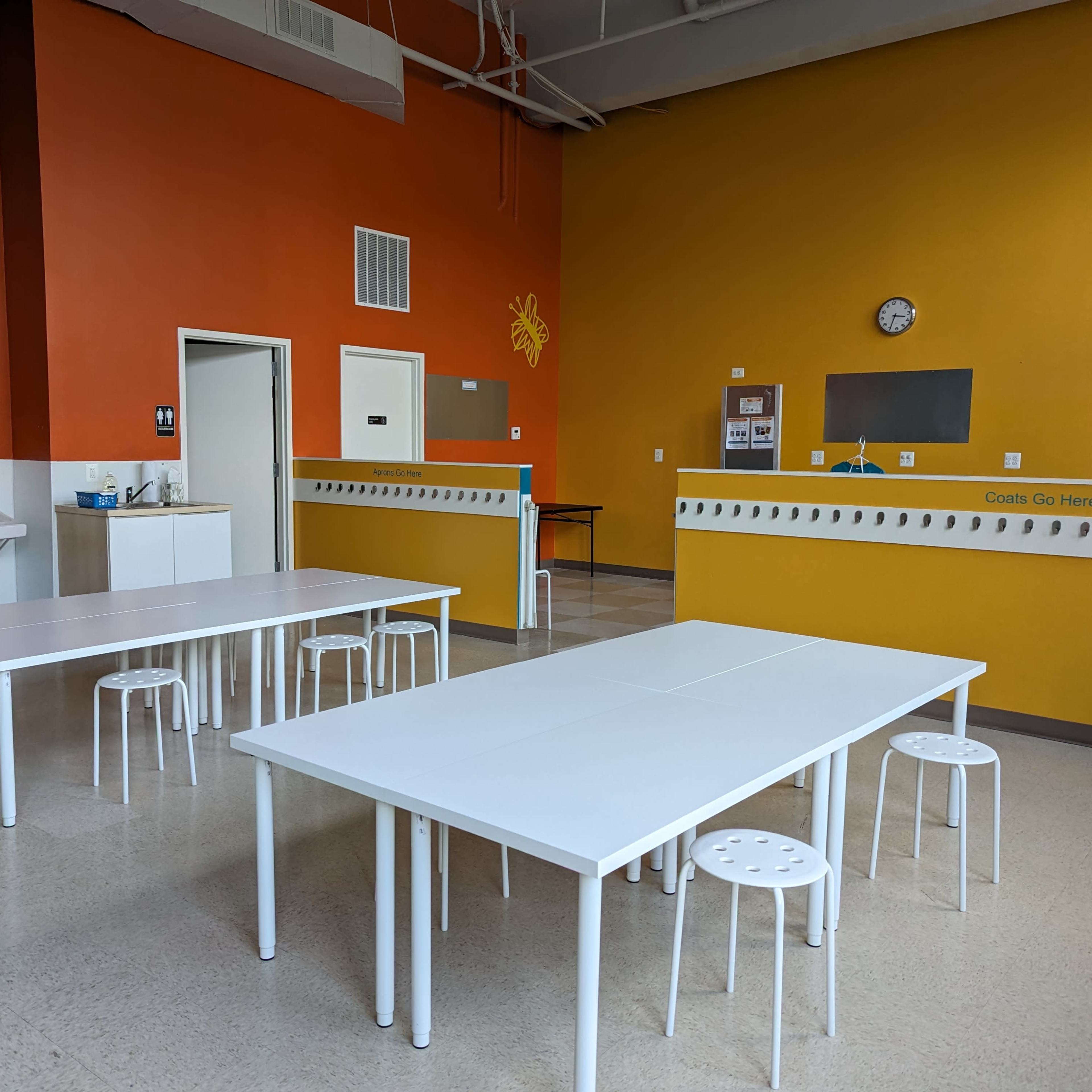 A spacious room with orange walls, two white tables, several white stools, and a sink area.