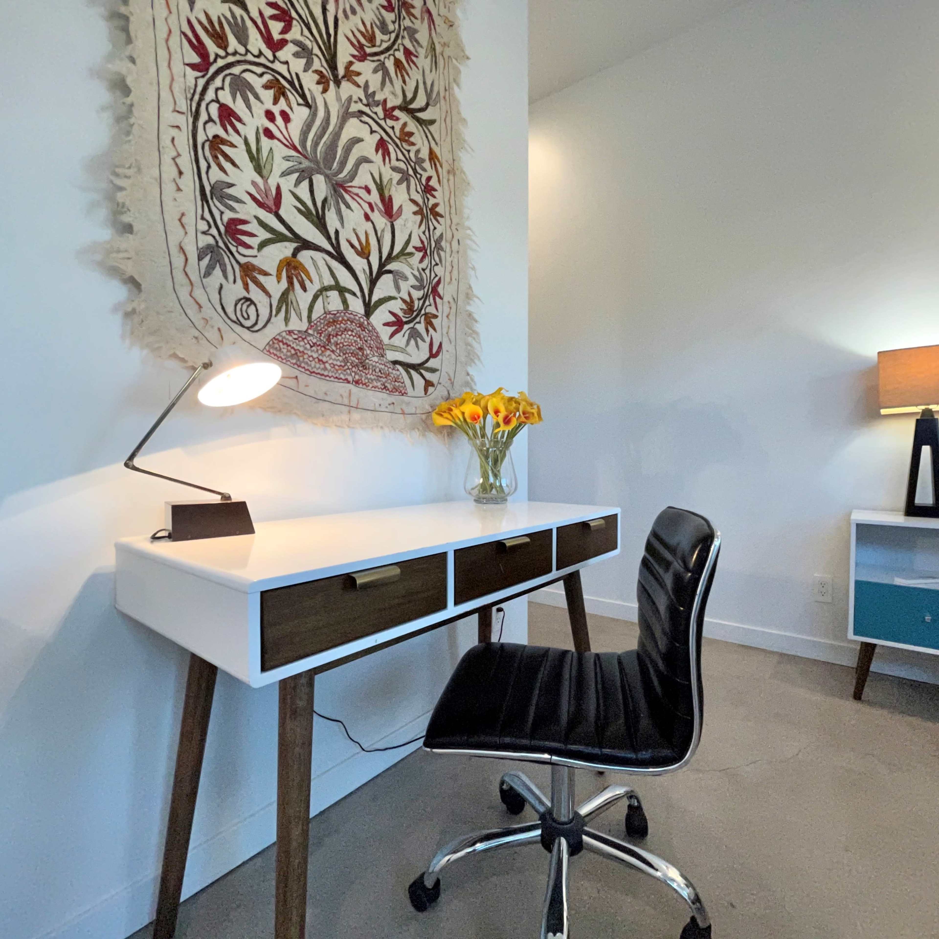 A modern workspace features a white desk with wooden accents, a black rolling chair, a lamp, a vase of yellow flowers, and a nightstand beside a bed.