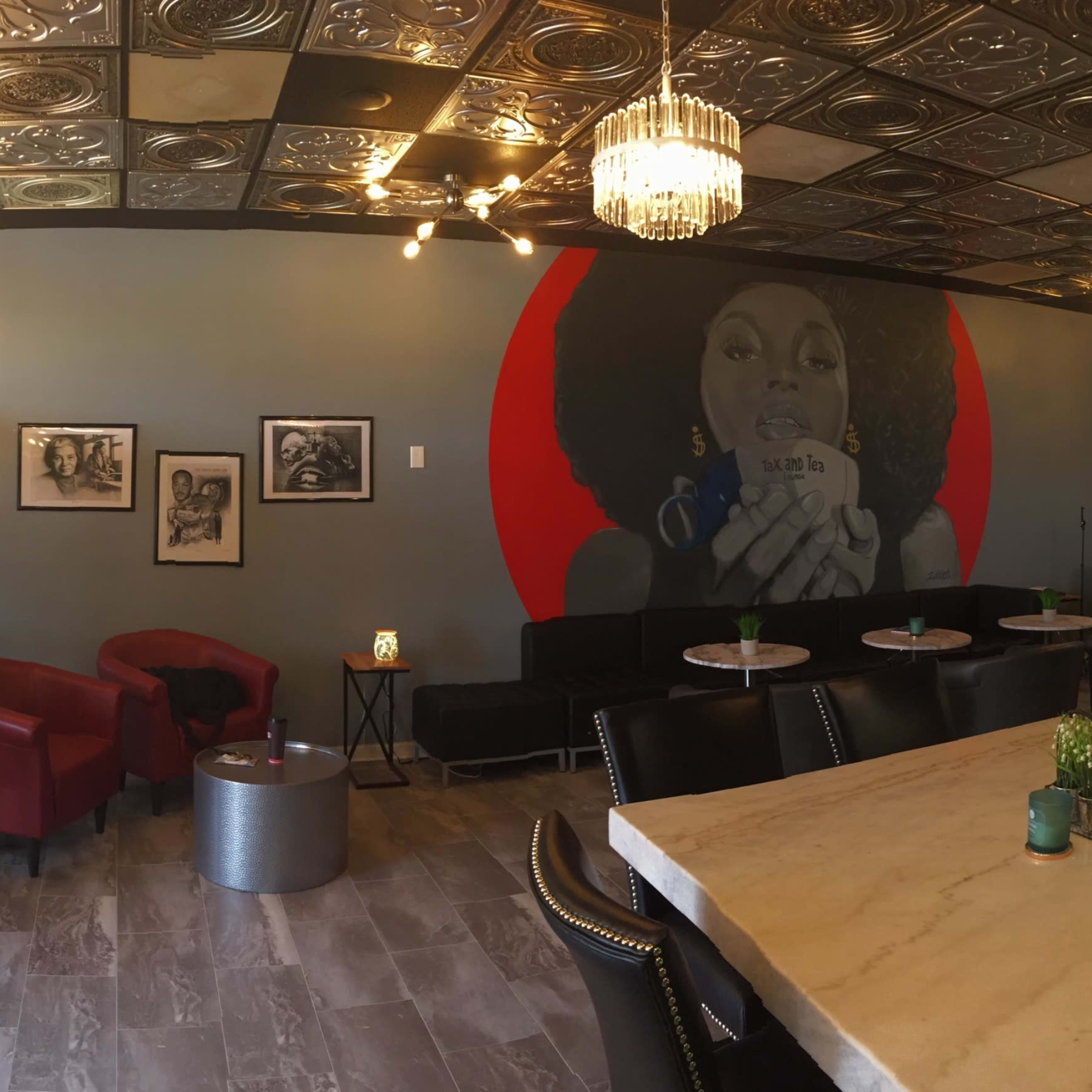 A modern café with a large mural on one wall, featuring a woman holding a cup, and a seating area consisting of a marble table and red armchairs.