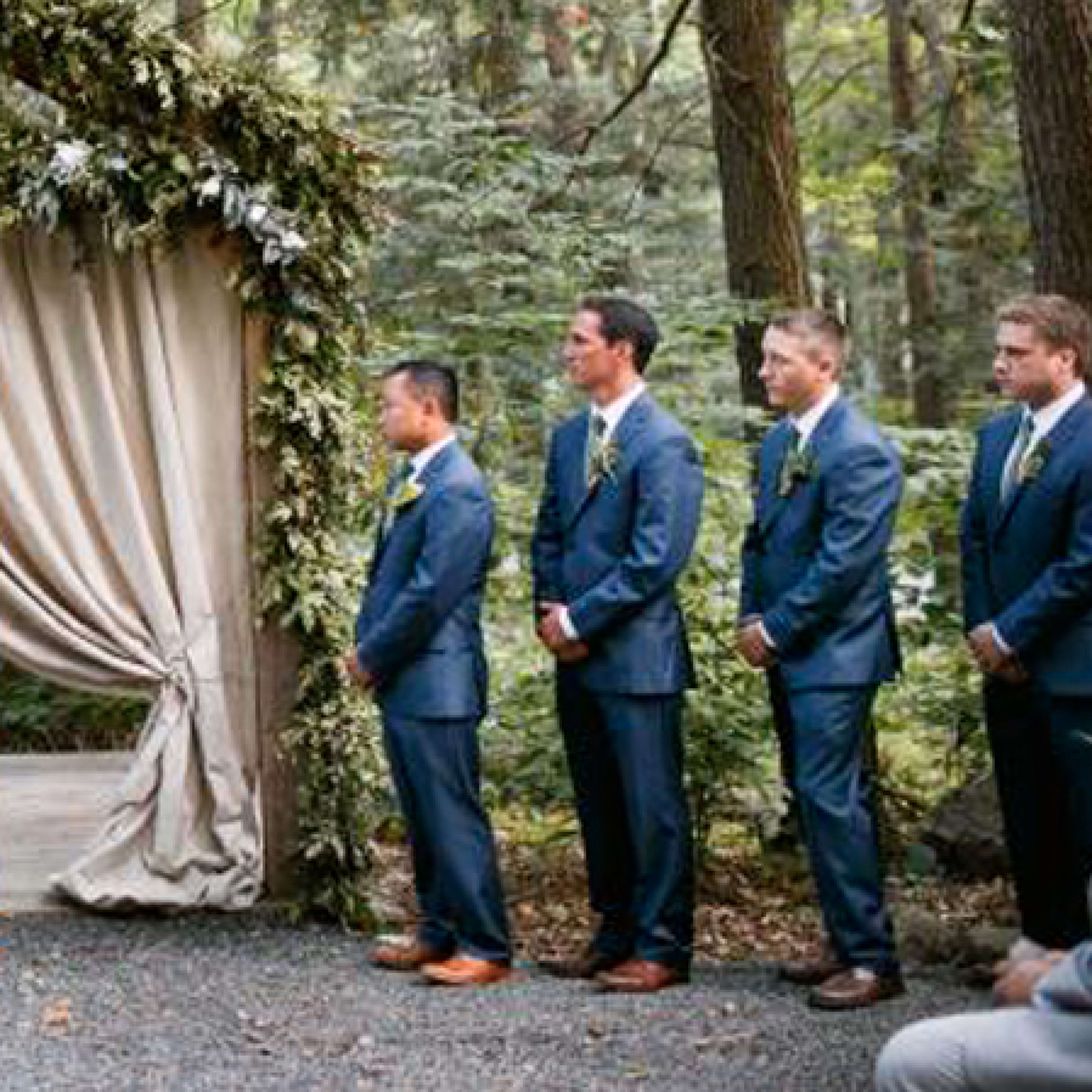 A wedding ceremony is taking place in a forested area under a floral arch, with the couple facing an officiant surrounded by their wedding party.