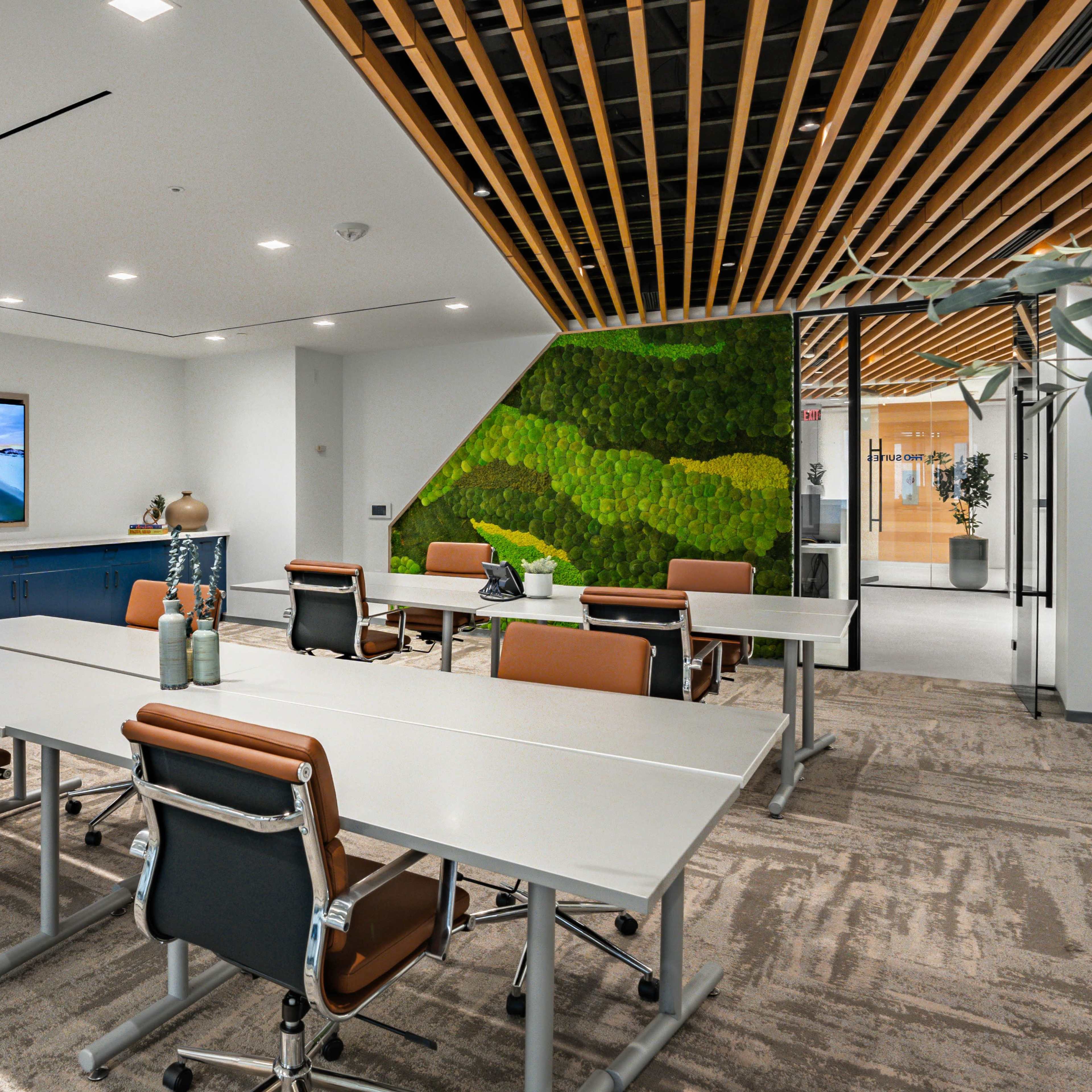 A modern conference room with several tables and chairs, a large television screen on the wall, and a green wall feature.