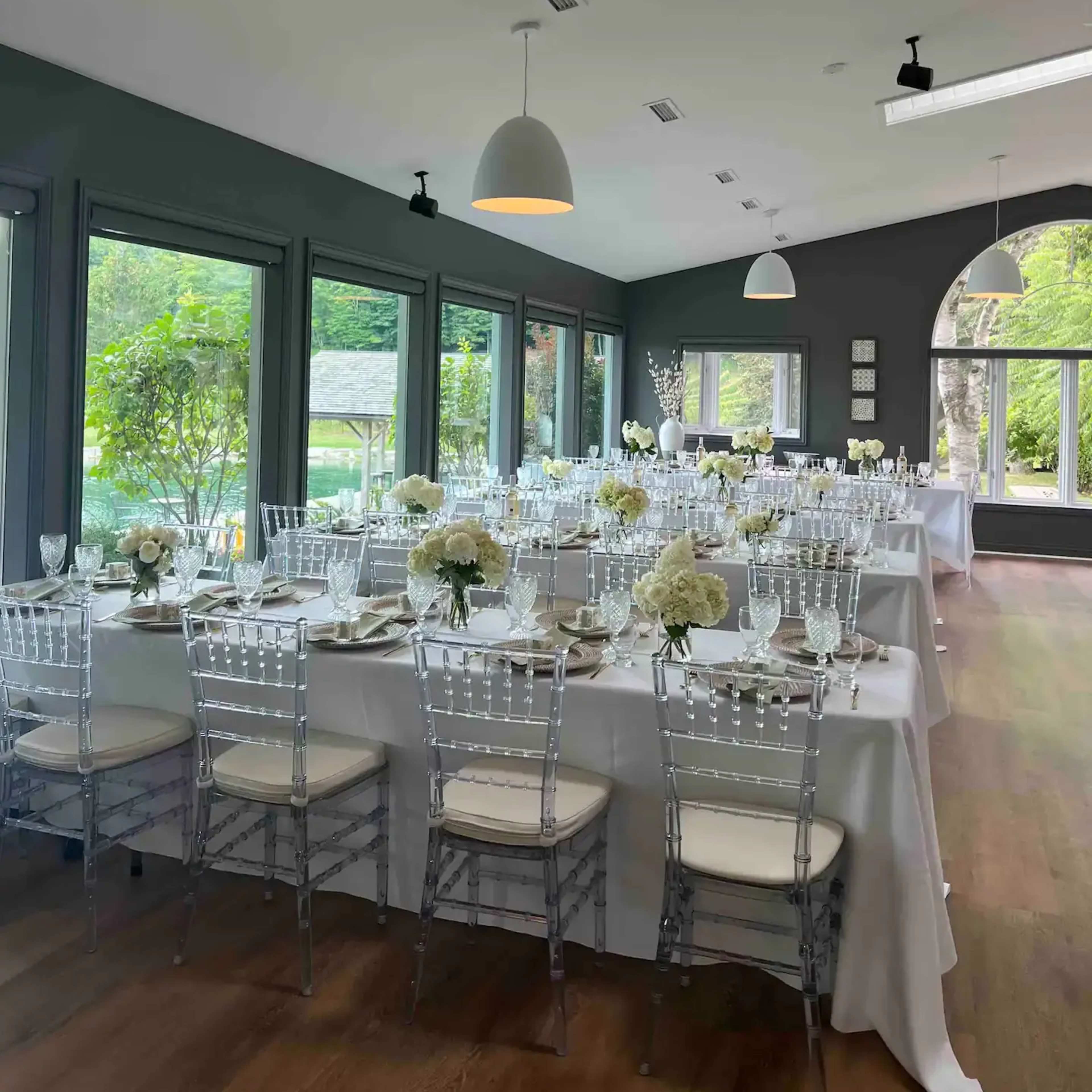 A dining area with elegant tables set for a meal, featuring white tablecloths and floral centerpieces, surrounded by large windows.