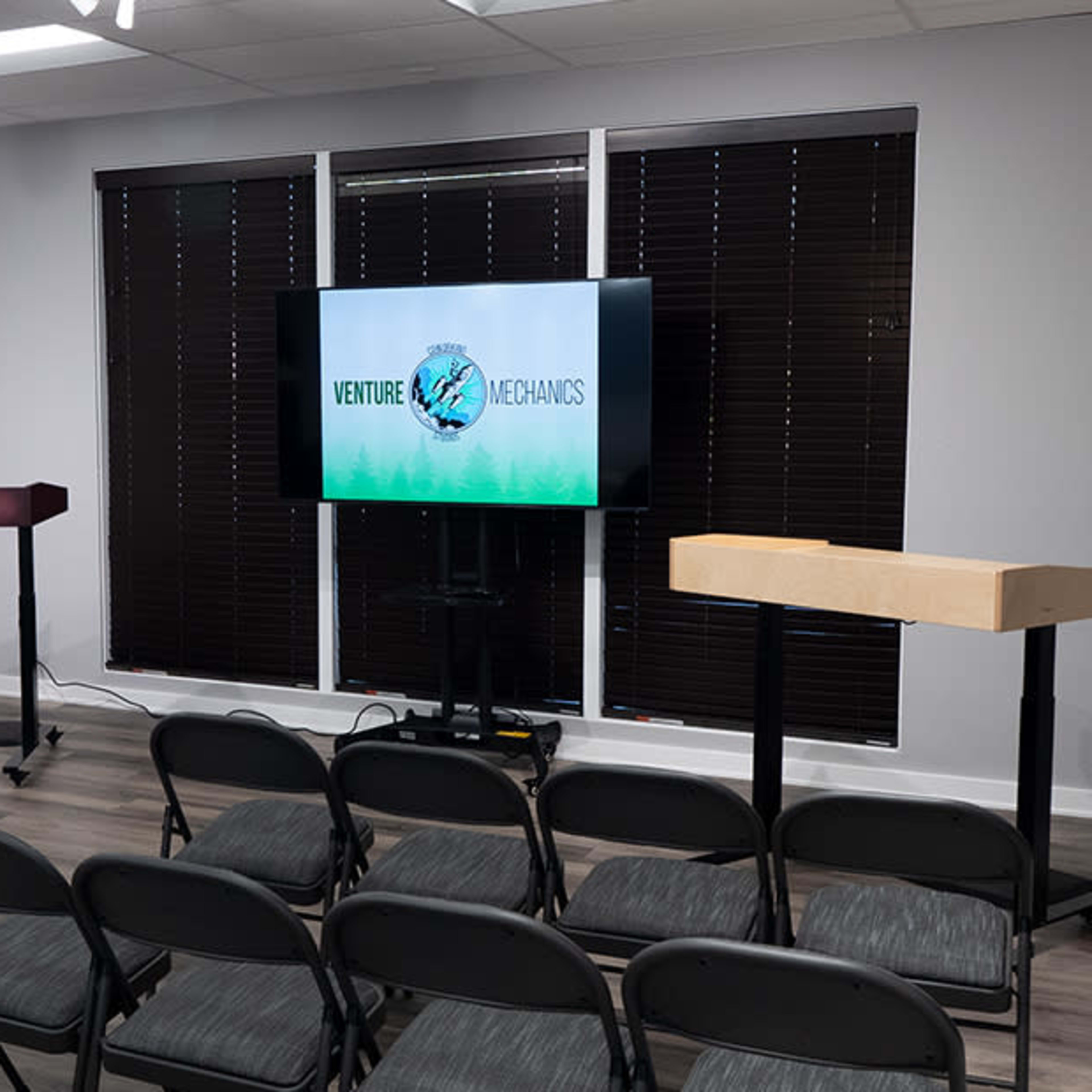 A conference room set up with black folding chairs facing two podiums and a large screen displaying the text "VENTURE METAMORPHOSIS."