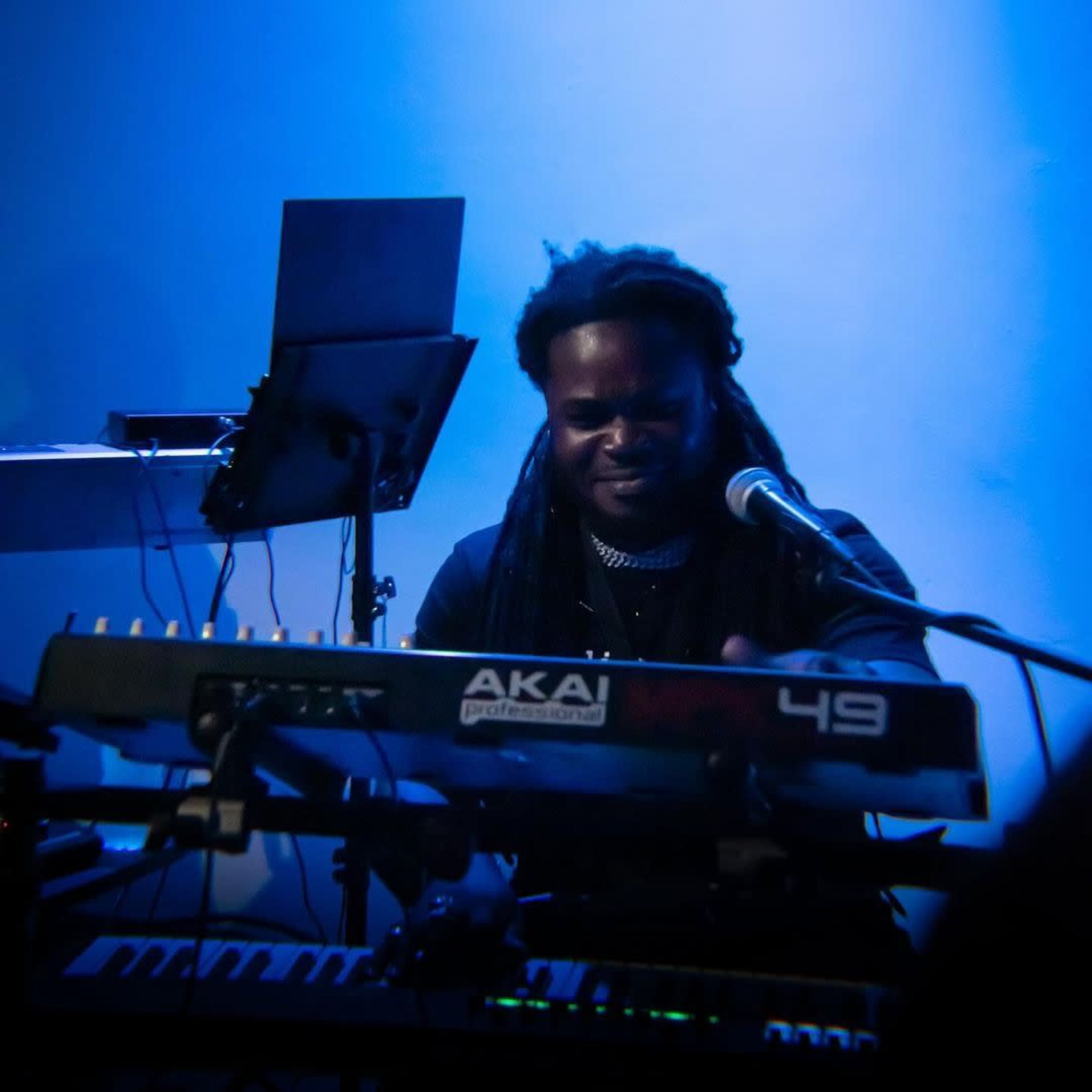 A musician plays an AKAI keyboard while singing into a microphone on stage.
