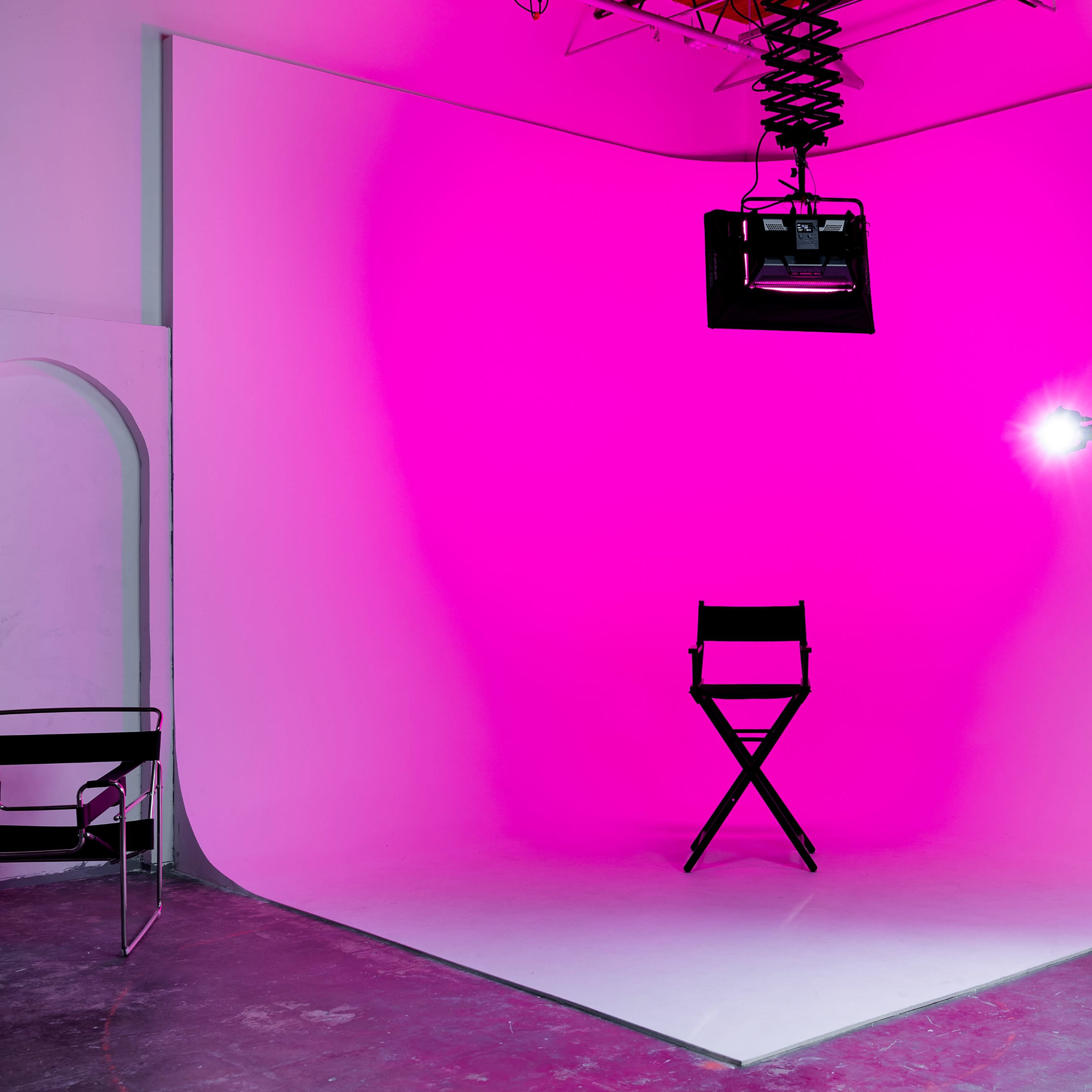 A photo studio setup featuring a black director's chair, a pink backdrop, a mounted light, and equipment on wheels.