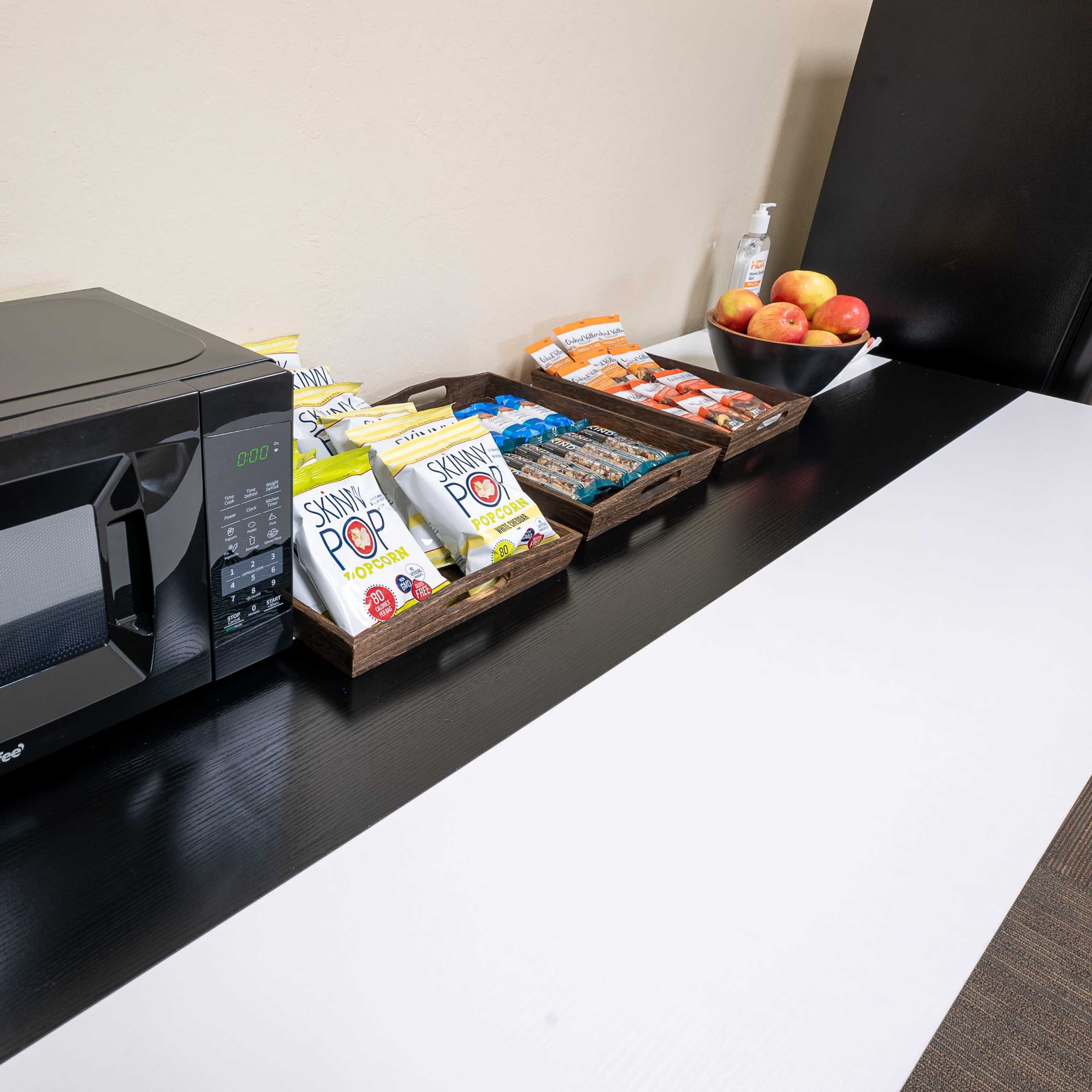 A black microwave is placed on a countertop next to various snacks and apples arranged in baskets.