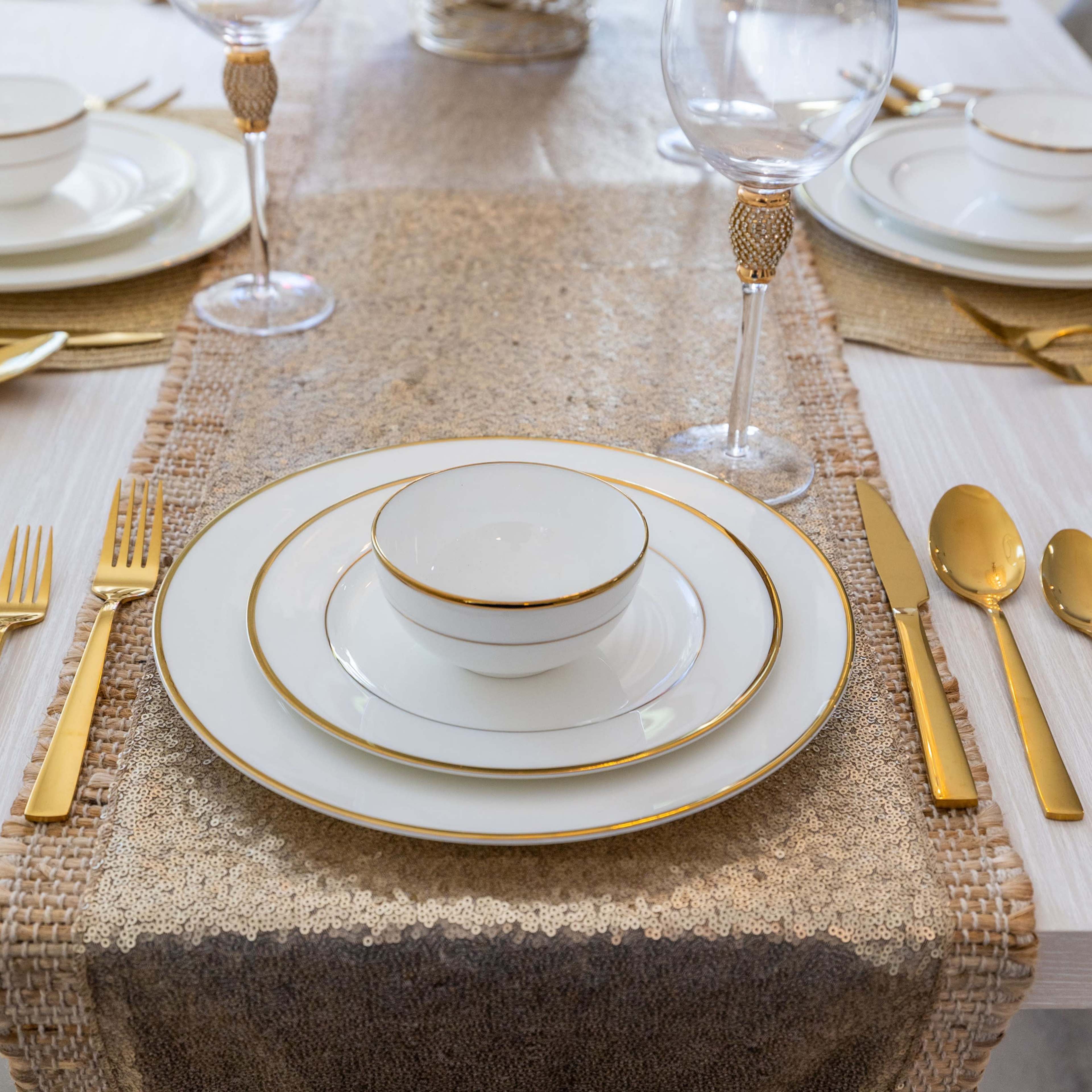 A dining table is elegantly set with white dishes featuring gold trim, sparkling silverware, and glassware on a decorative table runner.