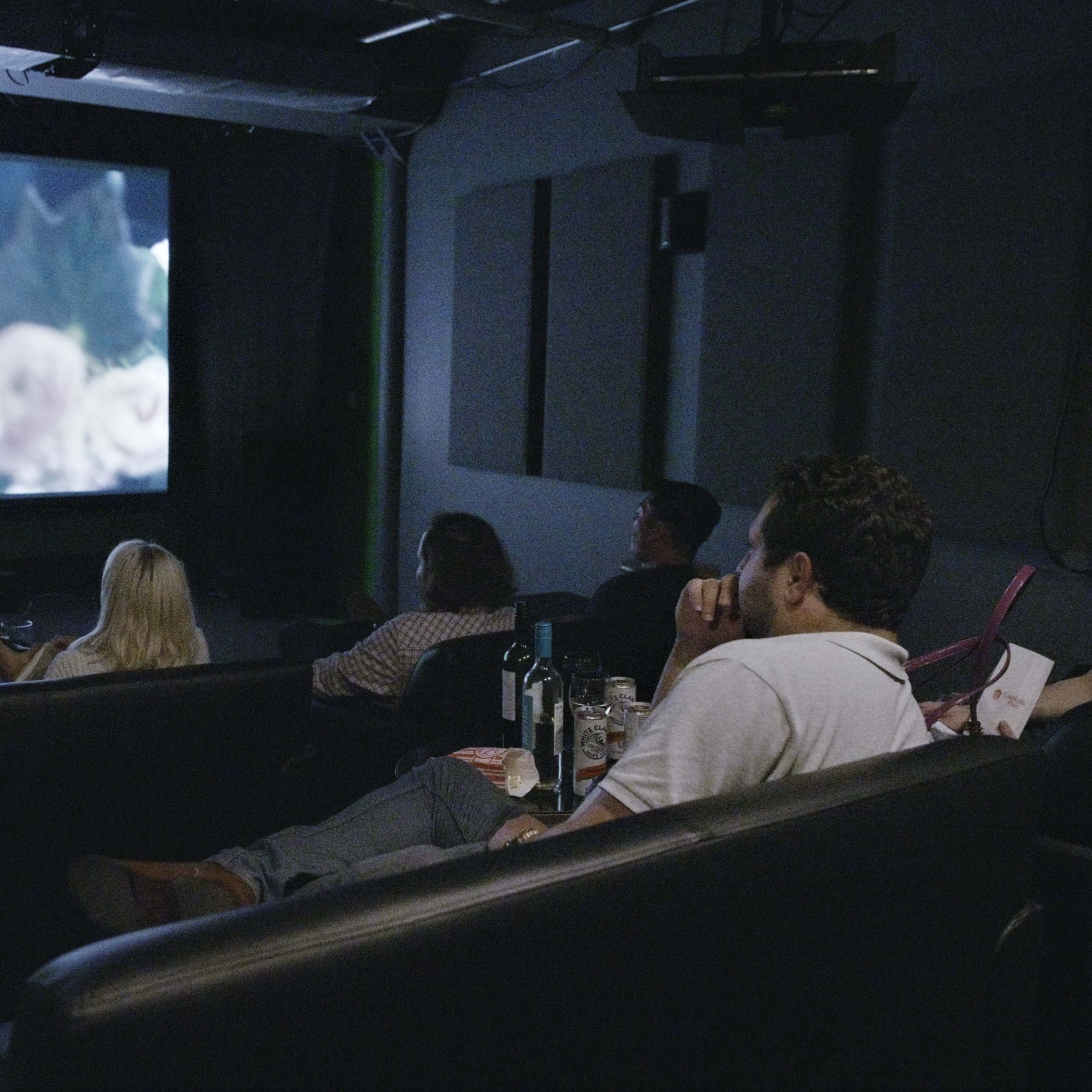 A group of people sits on a couch watching a movie on a large screen in a dimly lit room.