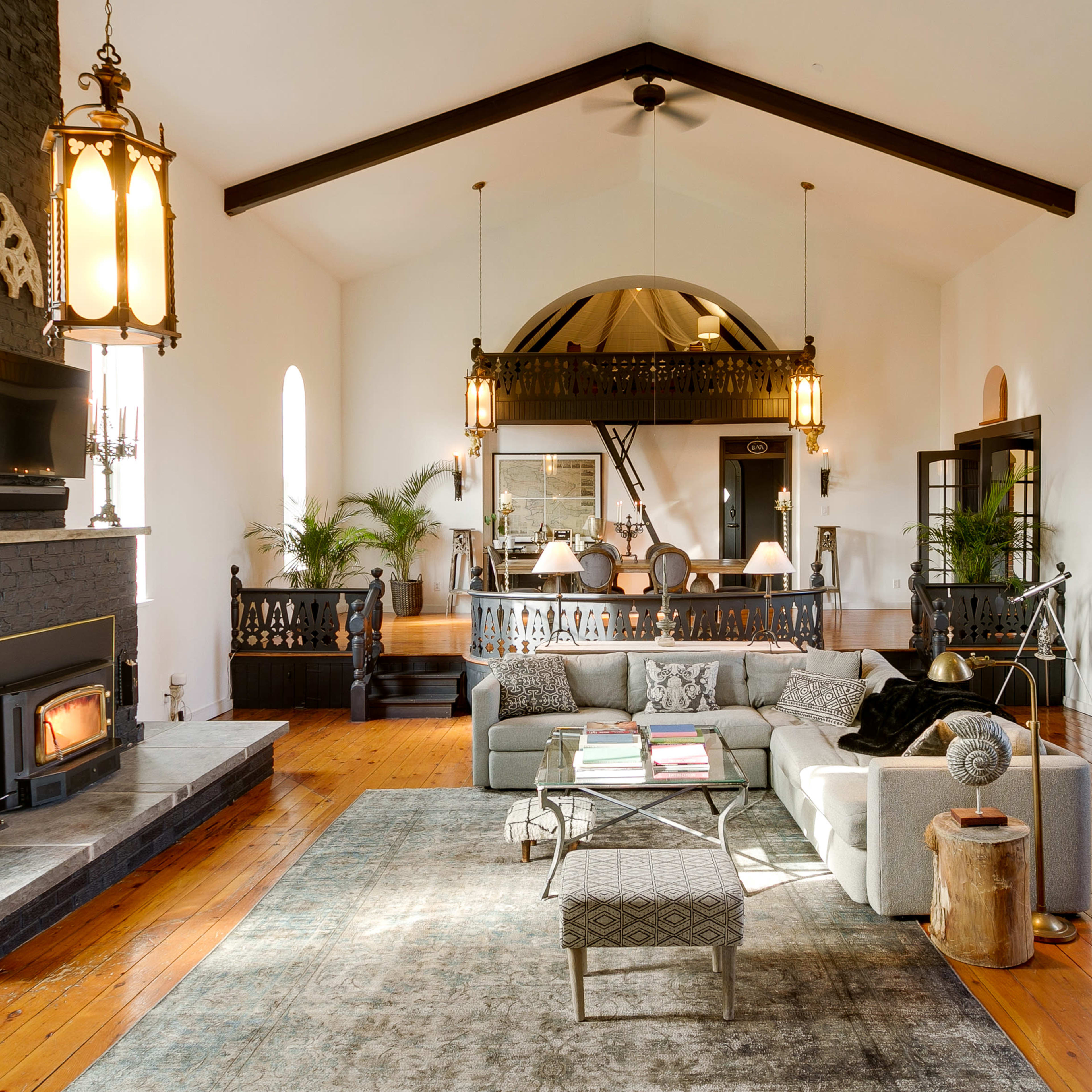 A spacious living room features high ceilings, large windows, a fireplace, and a mix of modern and traditional furnishings.