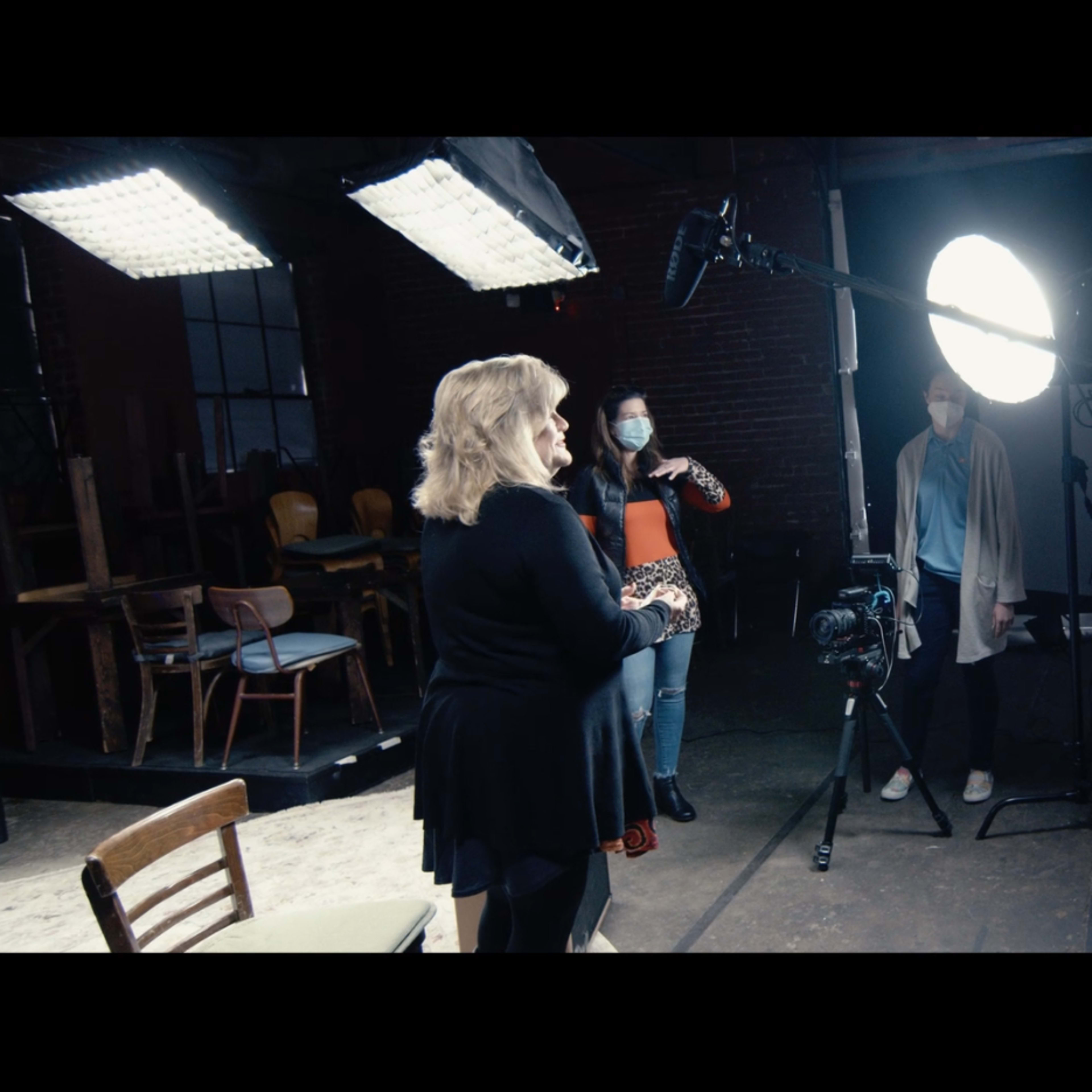 A small film crew sets up equipment in a studio, with a woman in front of a round light and cameras pointing towards her.
