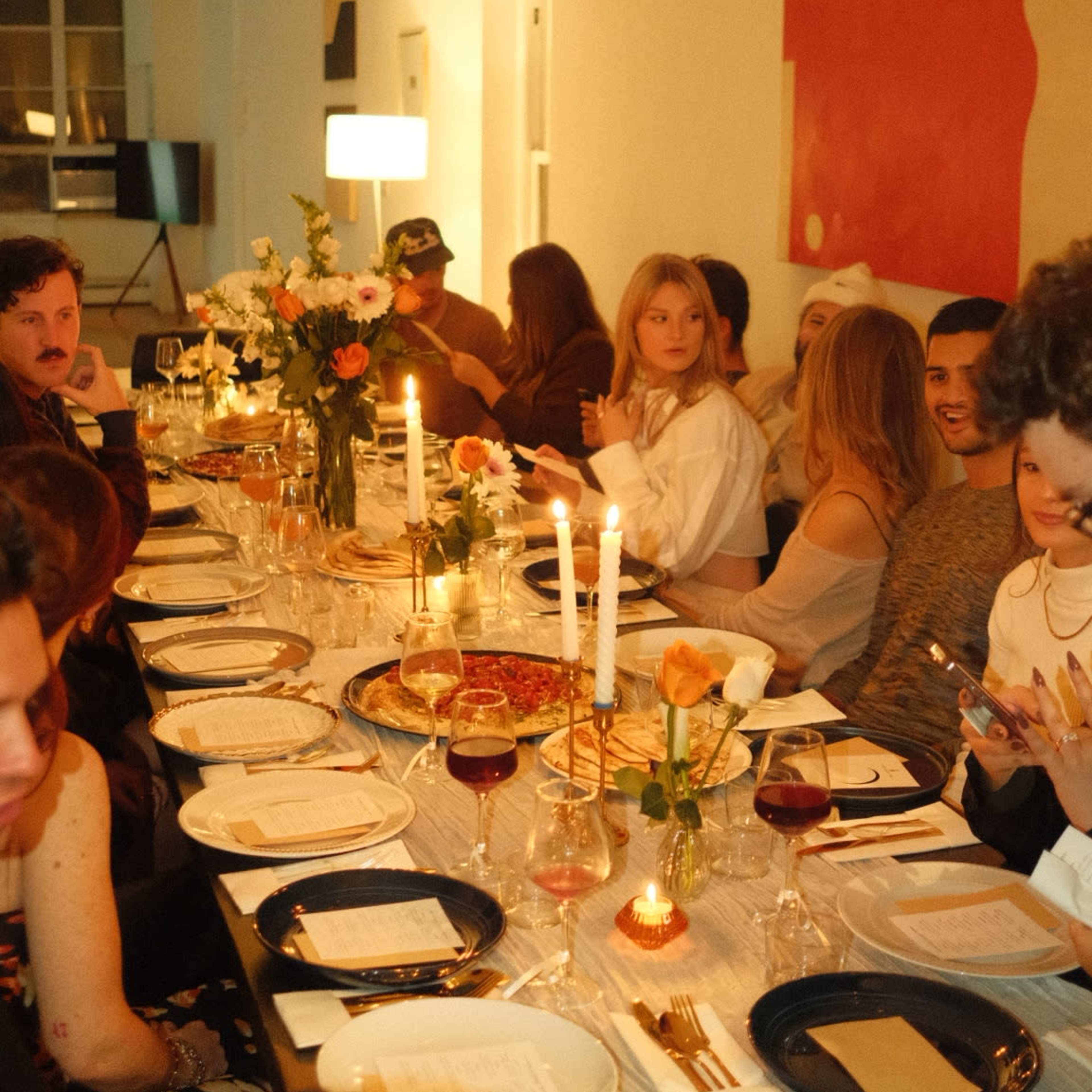 A long dining table is set with plates, glasses, and candles, as a group of people gather for a meal in a well-lit room decorated with artwork.
