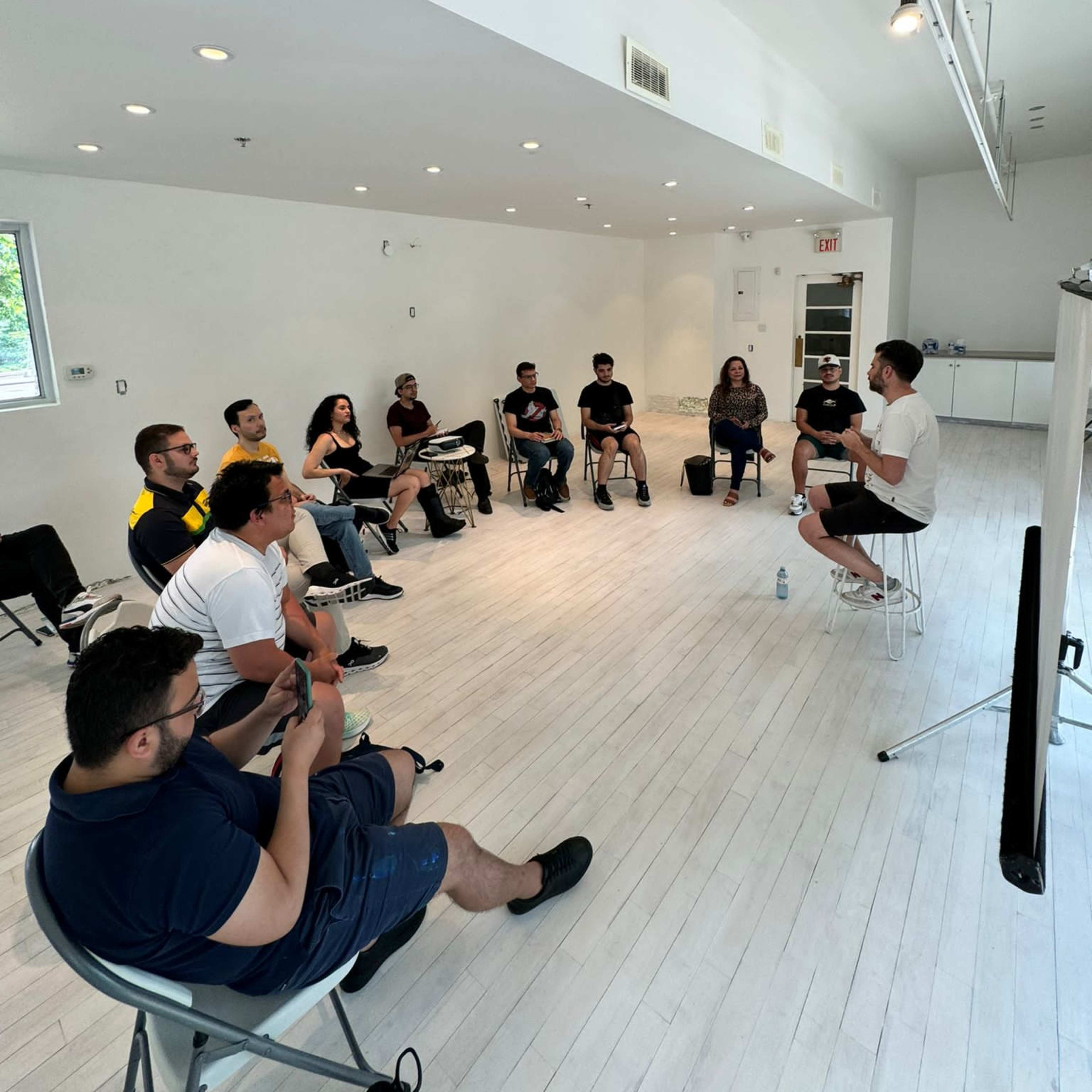 A group of people sits in a brightly lit room, facing a speaker who is standing and addressing them.