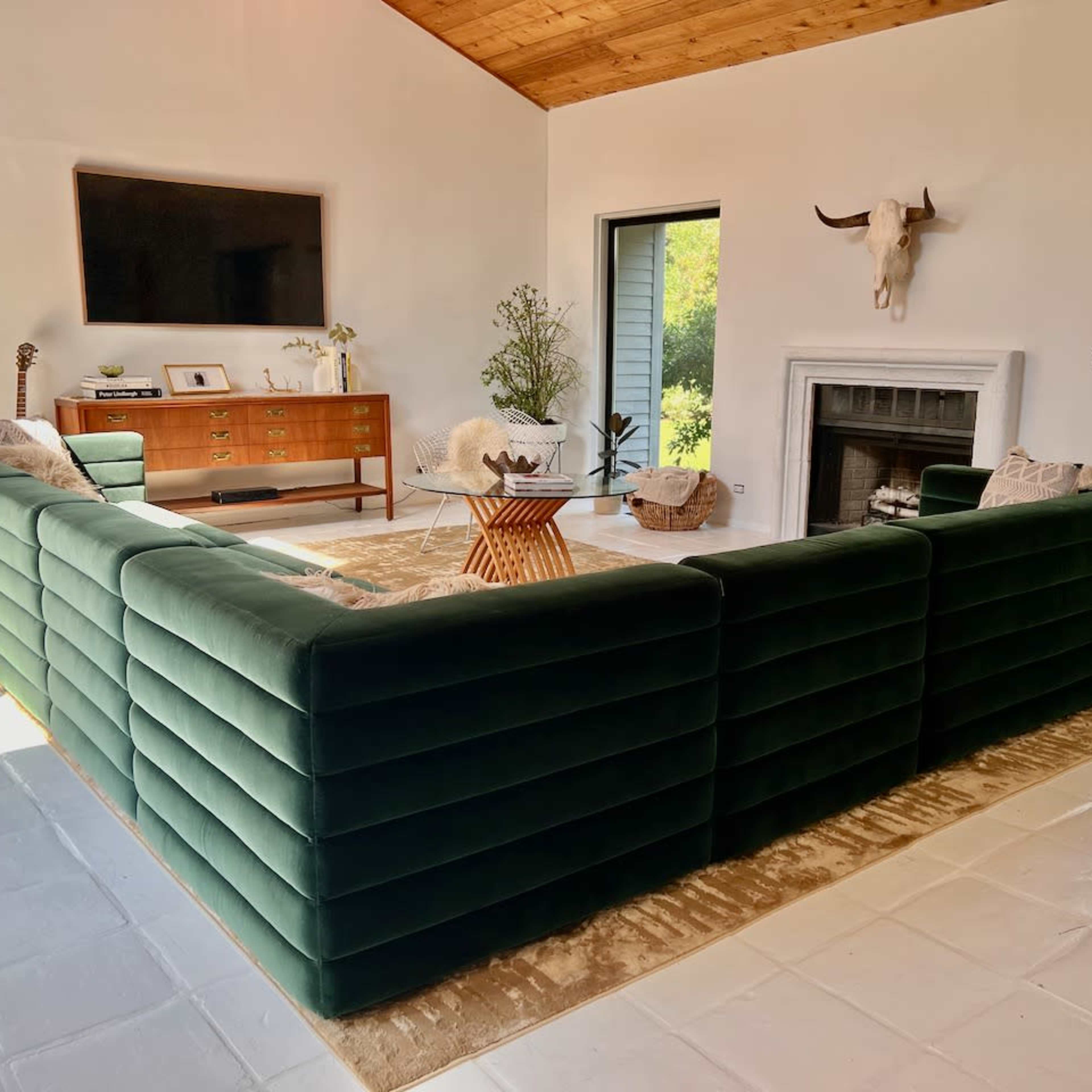 A modern living room features a large green velvet sectional sofa arranged around a central coffee table, with a television mounted on the wall and a fireplace nearby.