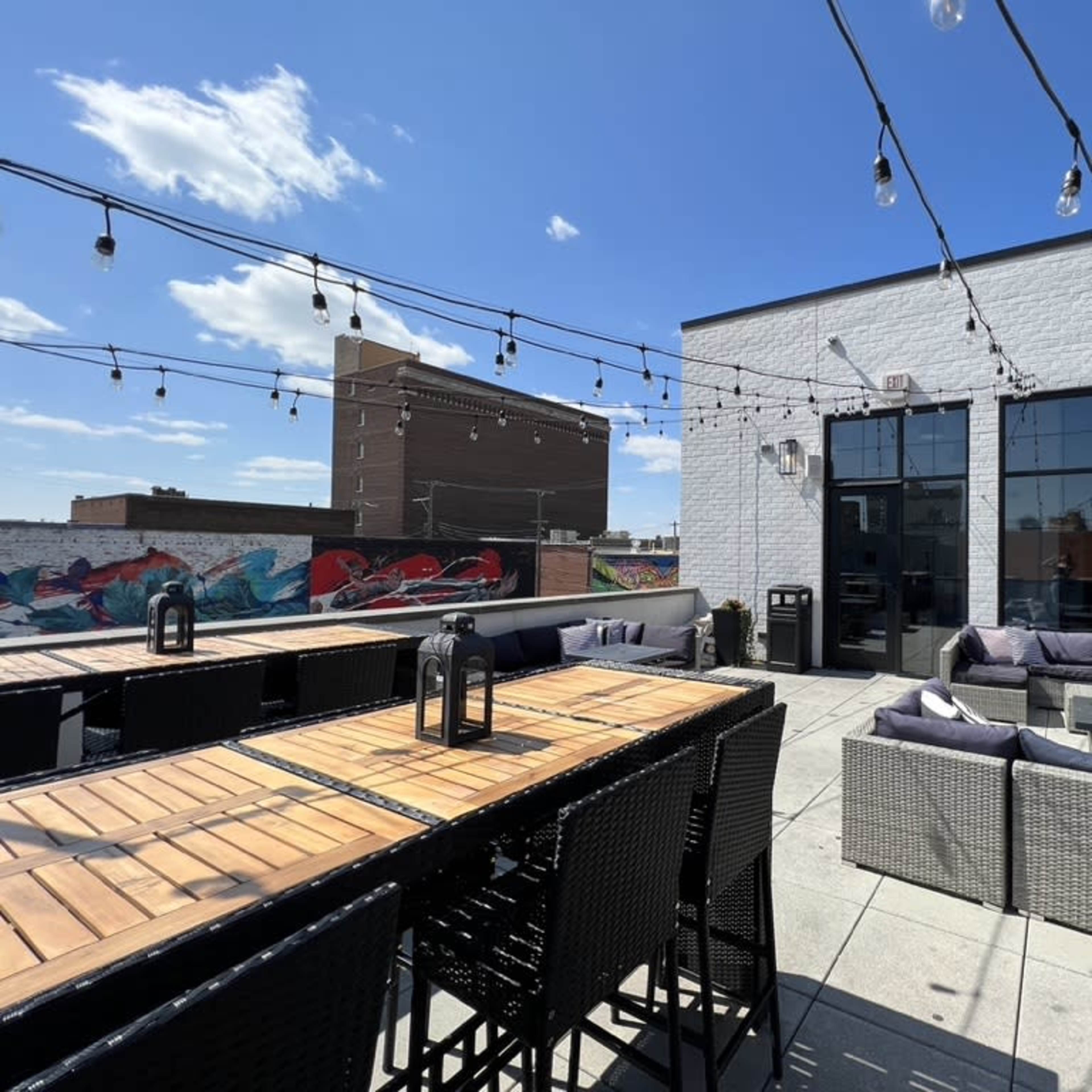 A rooftop patio features wooden tables, wicker seating, and string lights against a backdrop of a blue sky and city buildings.