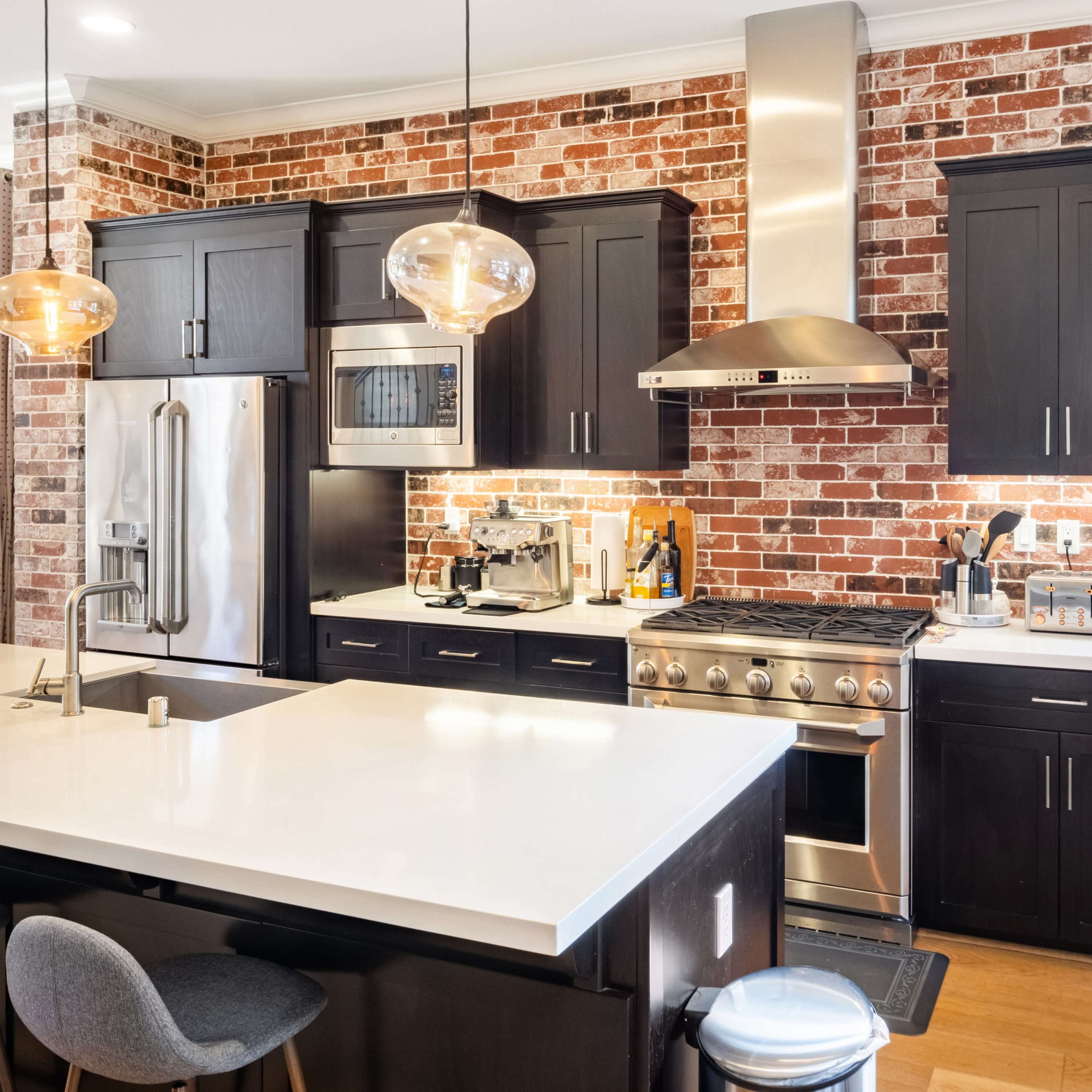 The kitchen features dark cabinetry, a stainless steel oven, and a large white island with pendant lighting, all set against a backdrop of exposed brick walls.