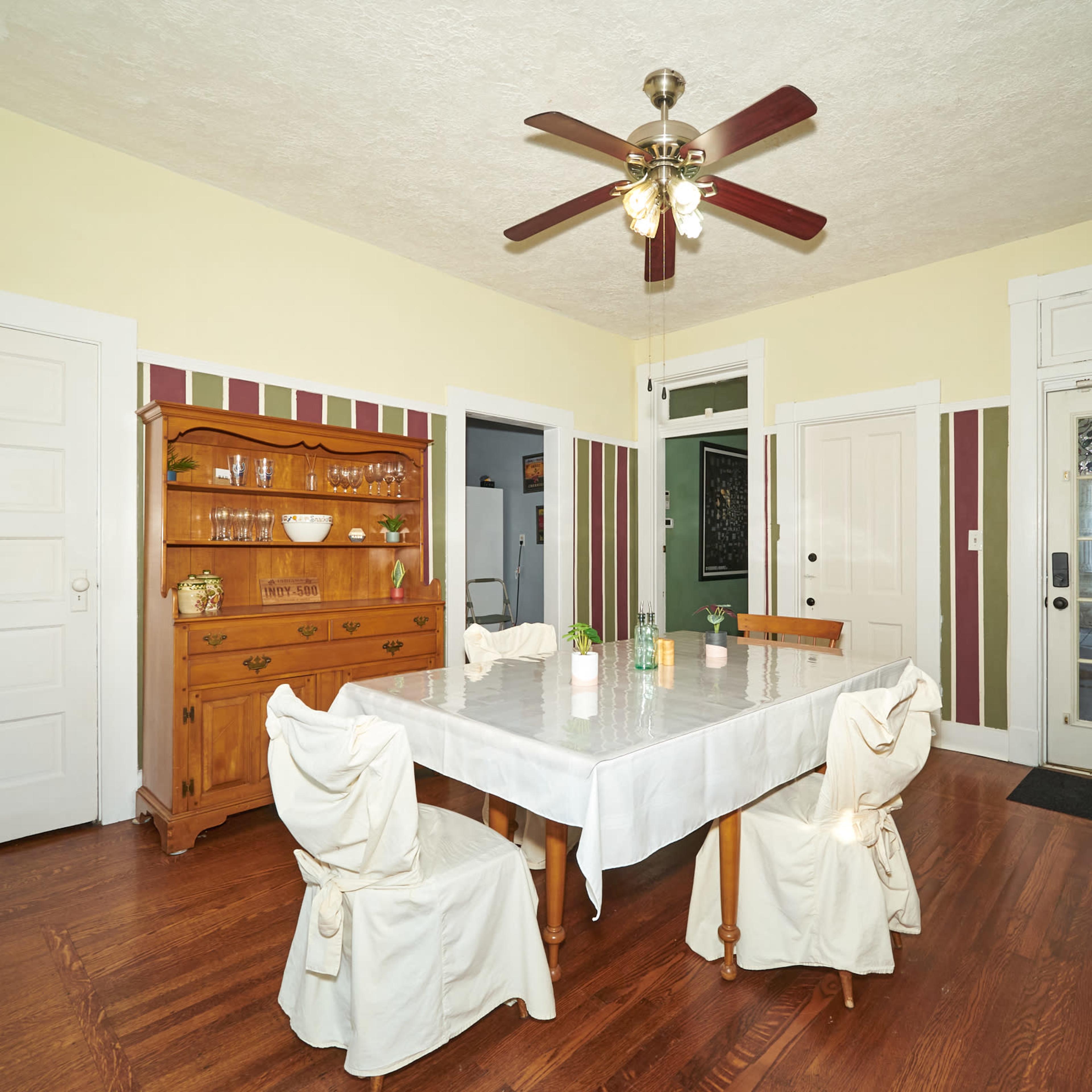 A dining room features a central table surrounded by six chairs, with a wooden sideboard and a ceiling fan above.