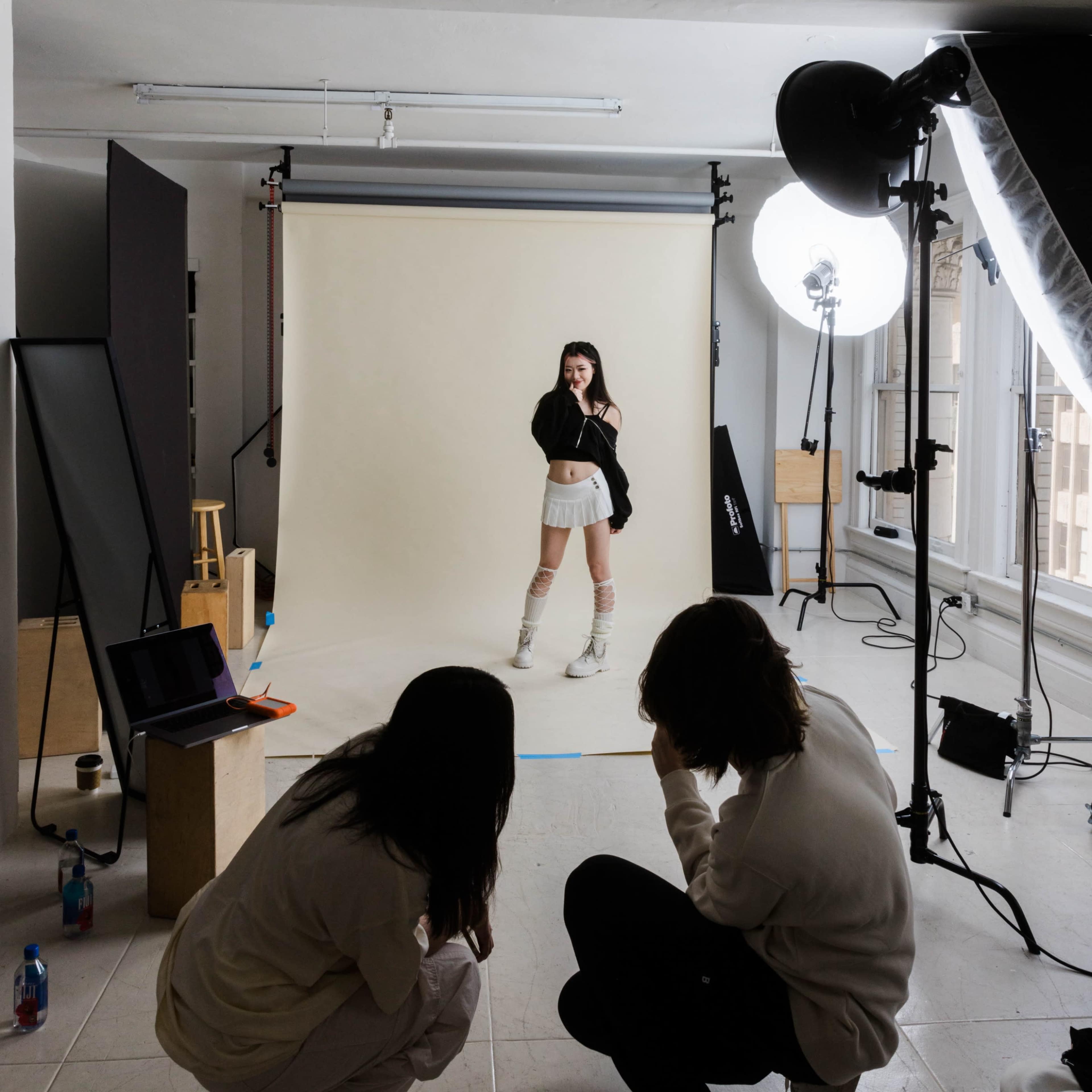 A model poses in front of a neutral backdrop while two photographers observe and adjust their equipment in a studio.