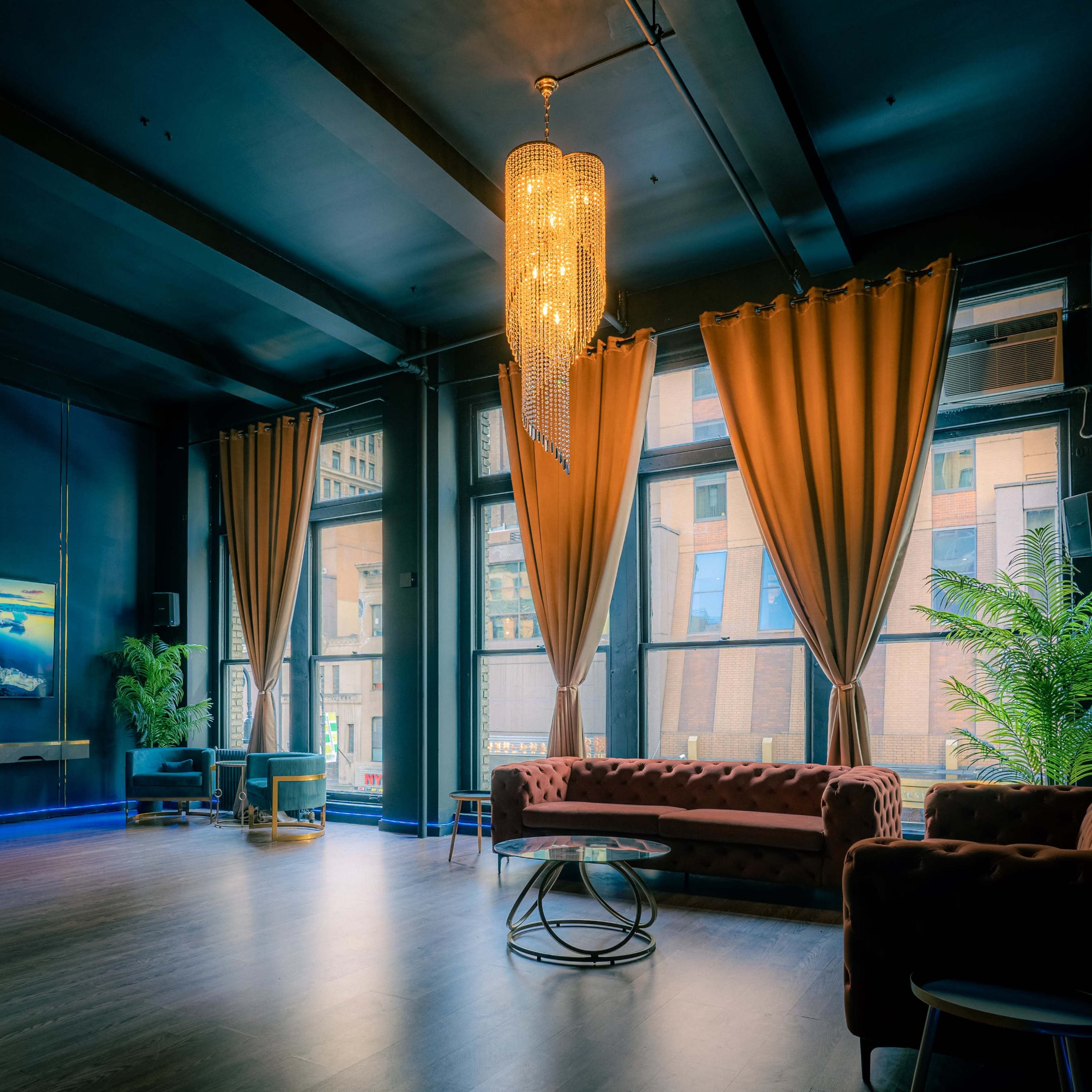 A spacious room features large windows with drapes, a chandelier, a sofa, and potted plants.