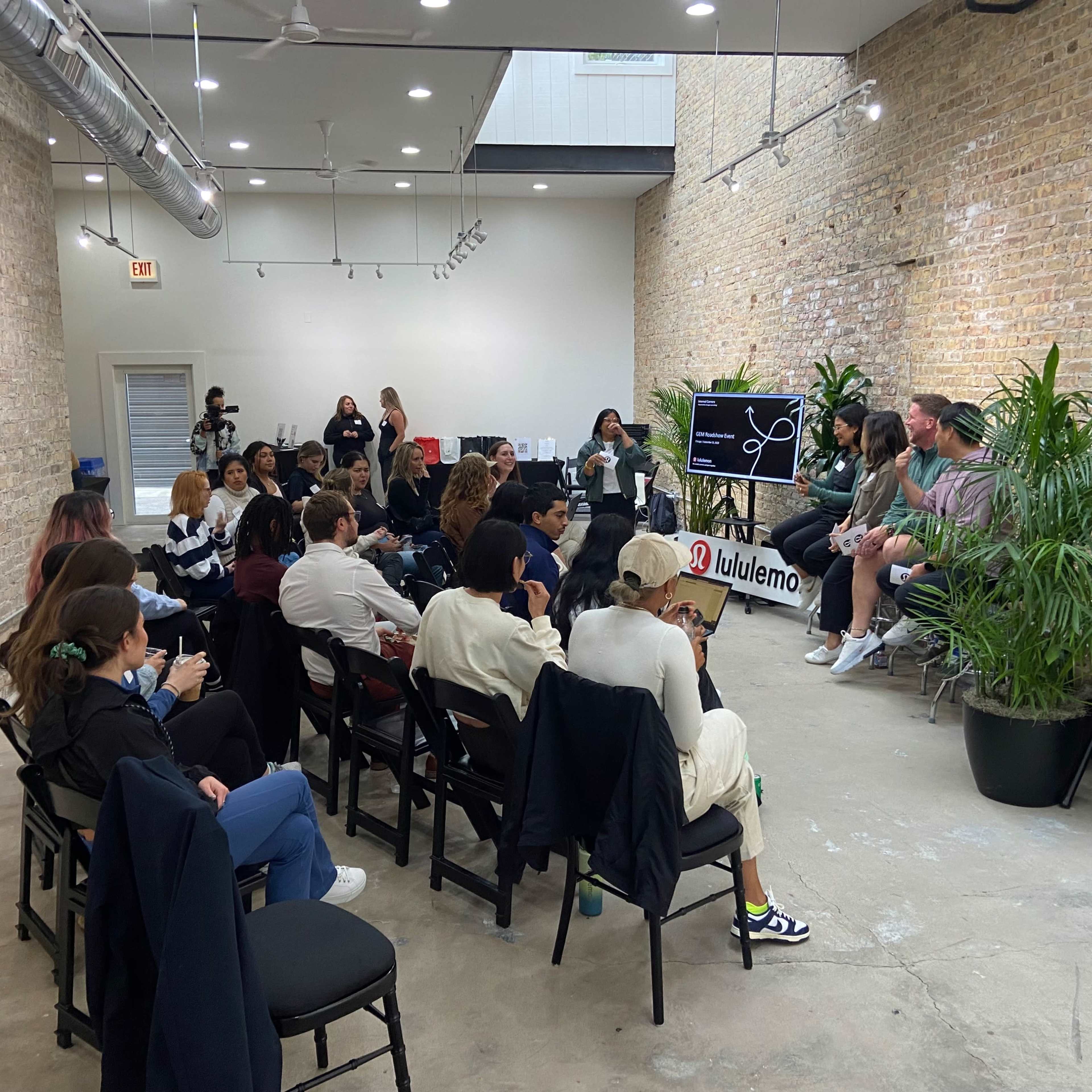 A group of people sits in chairs facing a panel discussion in a spacious room with a brick wall and plant decorations.