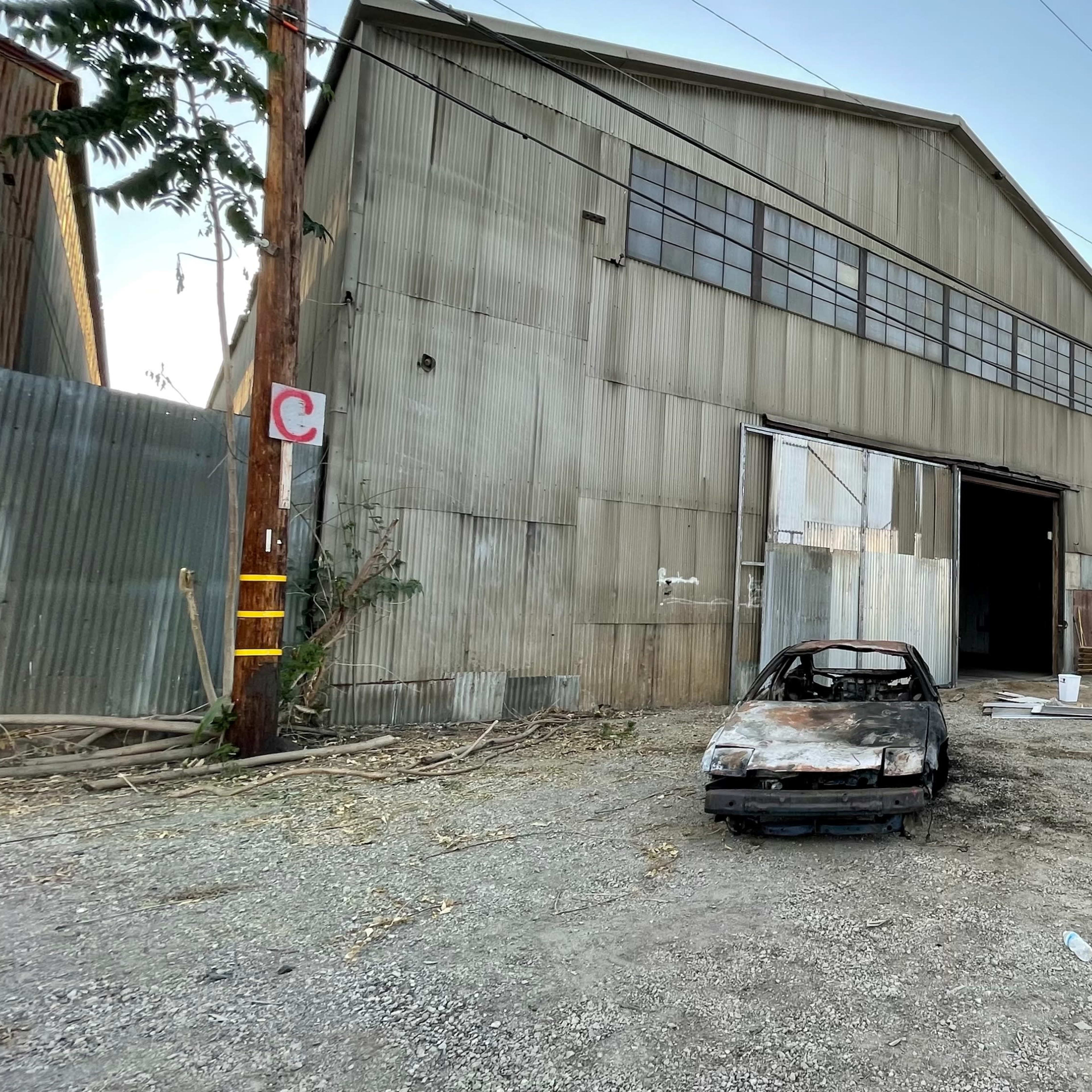 An old, burned-out car sits in front of a large, corrugated metal warehouse in a gravel lot.