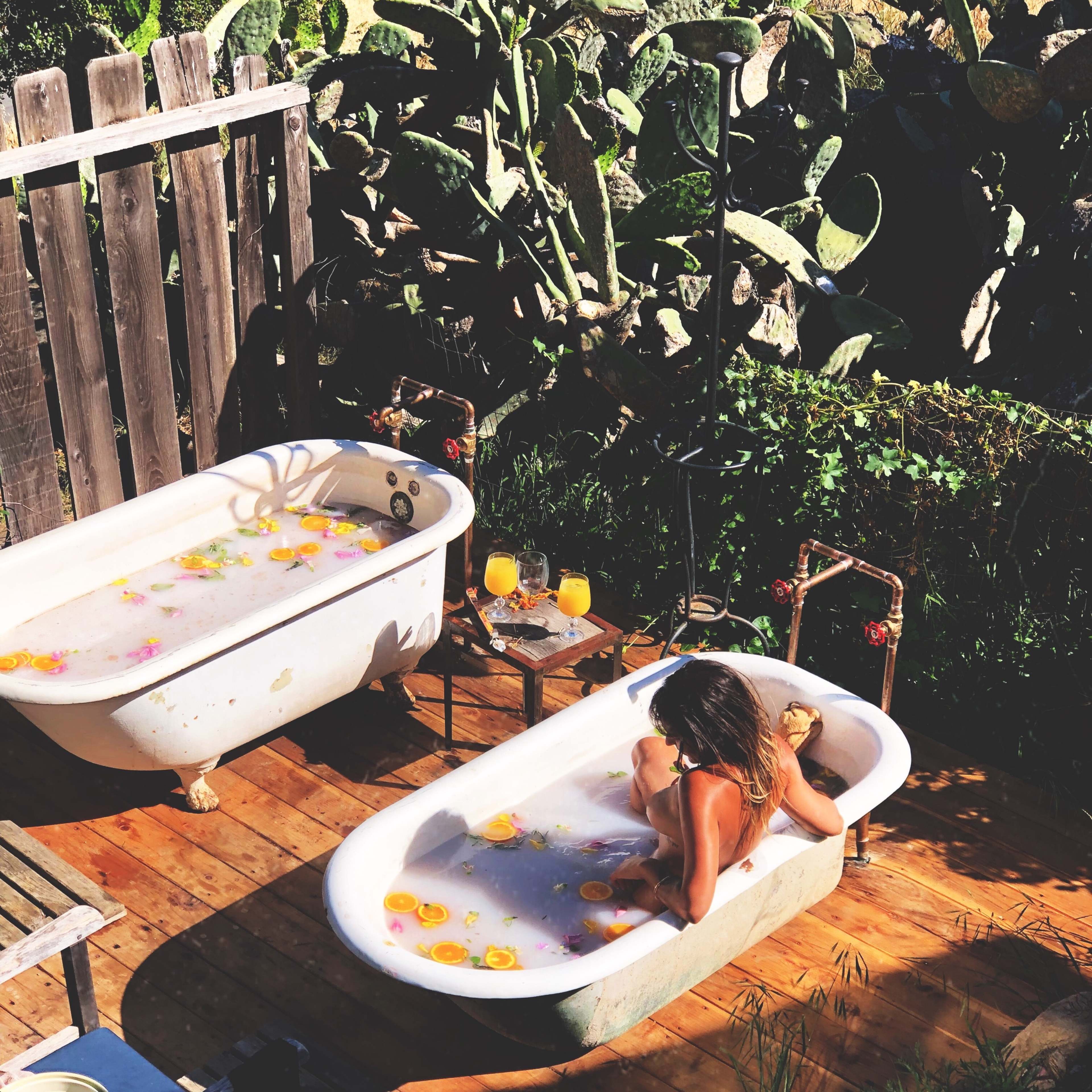 Two vintage bathtubs filled with water and flower petals sit on a wooden deck surrounded by cacti, while a person relaxes in one of the tubs.