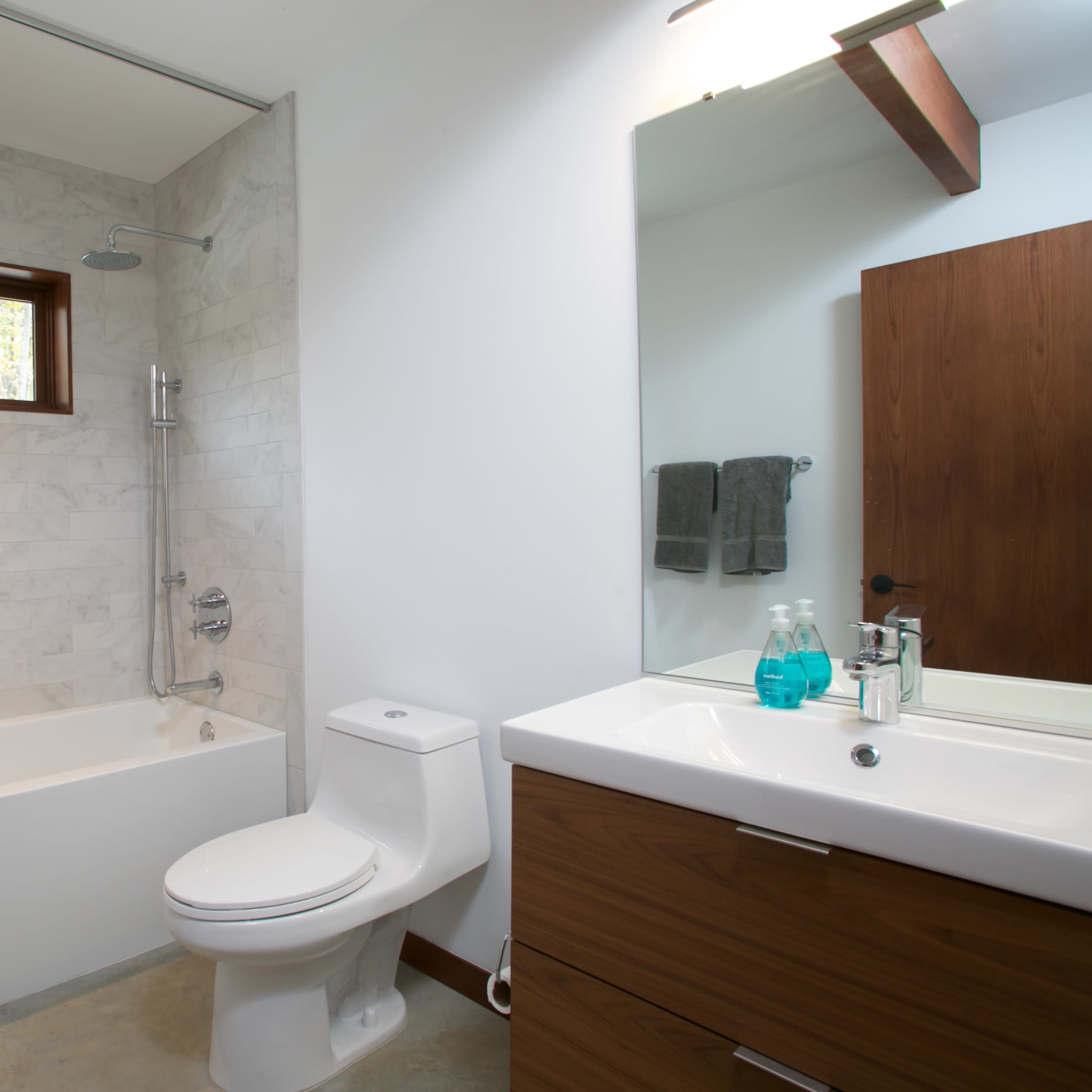 A modern bathroom featuring a bathtub with a shower, a wall-mounted mirror, a single vanity with drawers, and a toilet, all set against a backdrop of light-colored tiles and a wooden door.