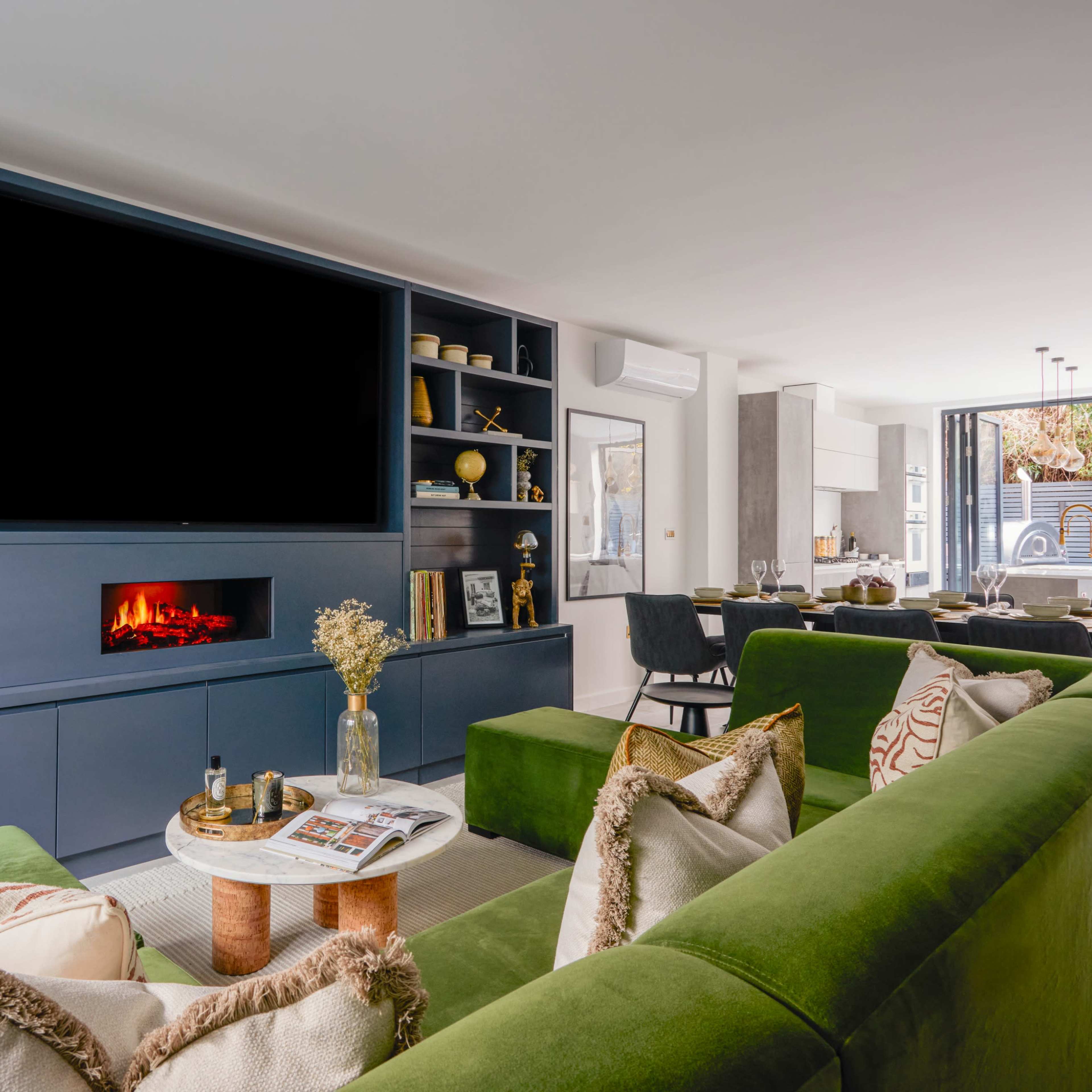 A modern living room features a large dark blue media unit with an empty television space, a coffee table with magazines, and green sofas arranged in a comfortable seating area.