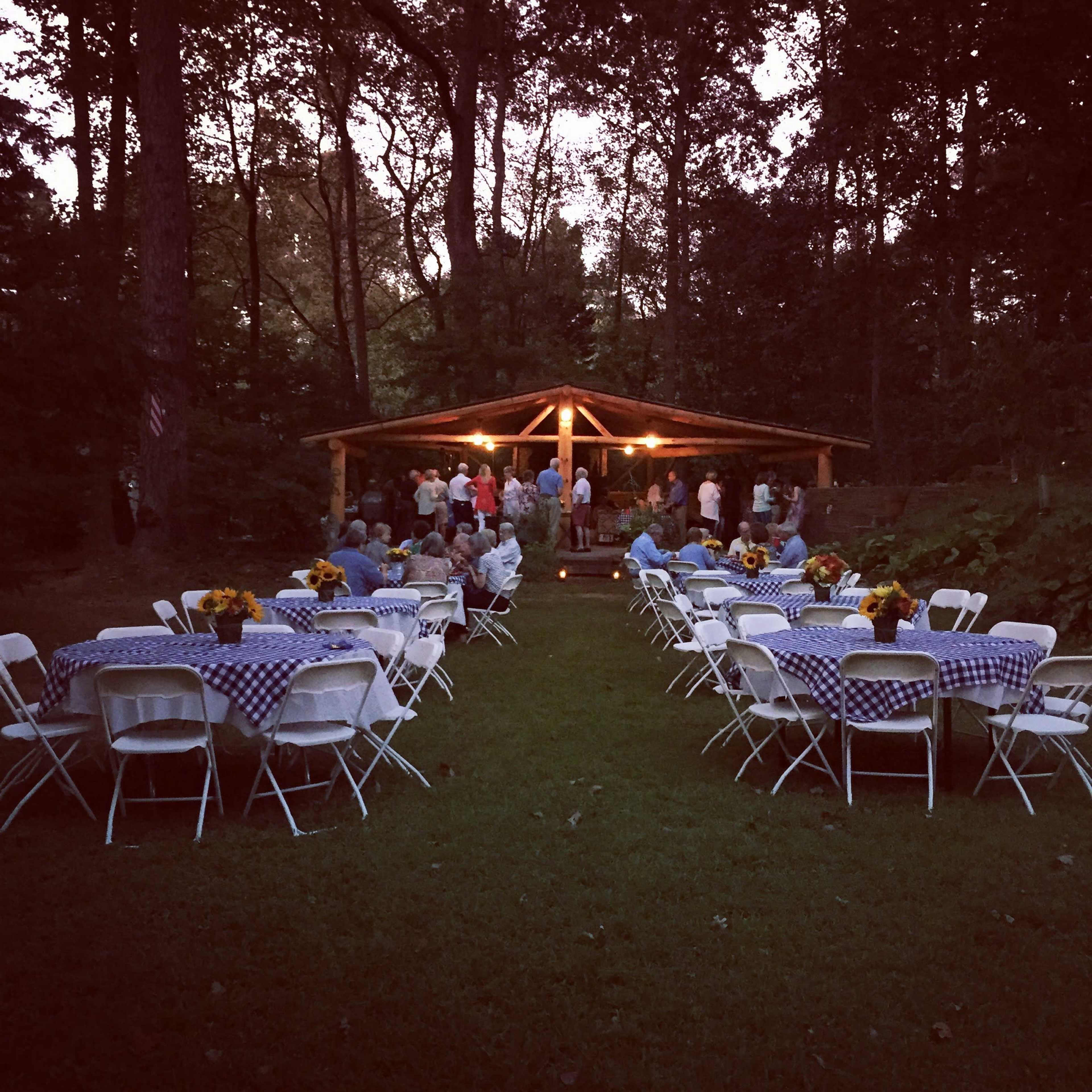 A gathering of people under a pavilion in a wooded area, with tables arranged in rows covered in blue and white checkered cloths.