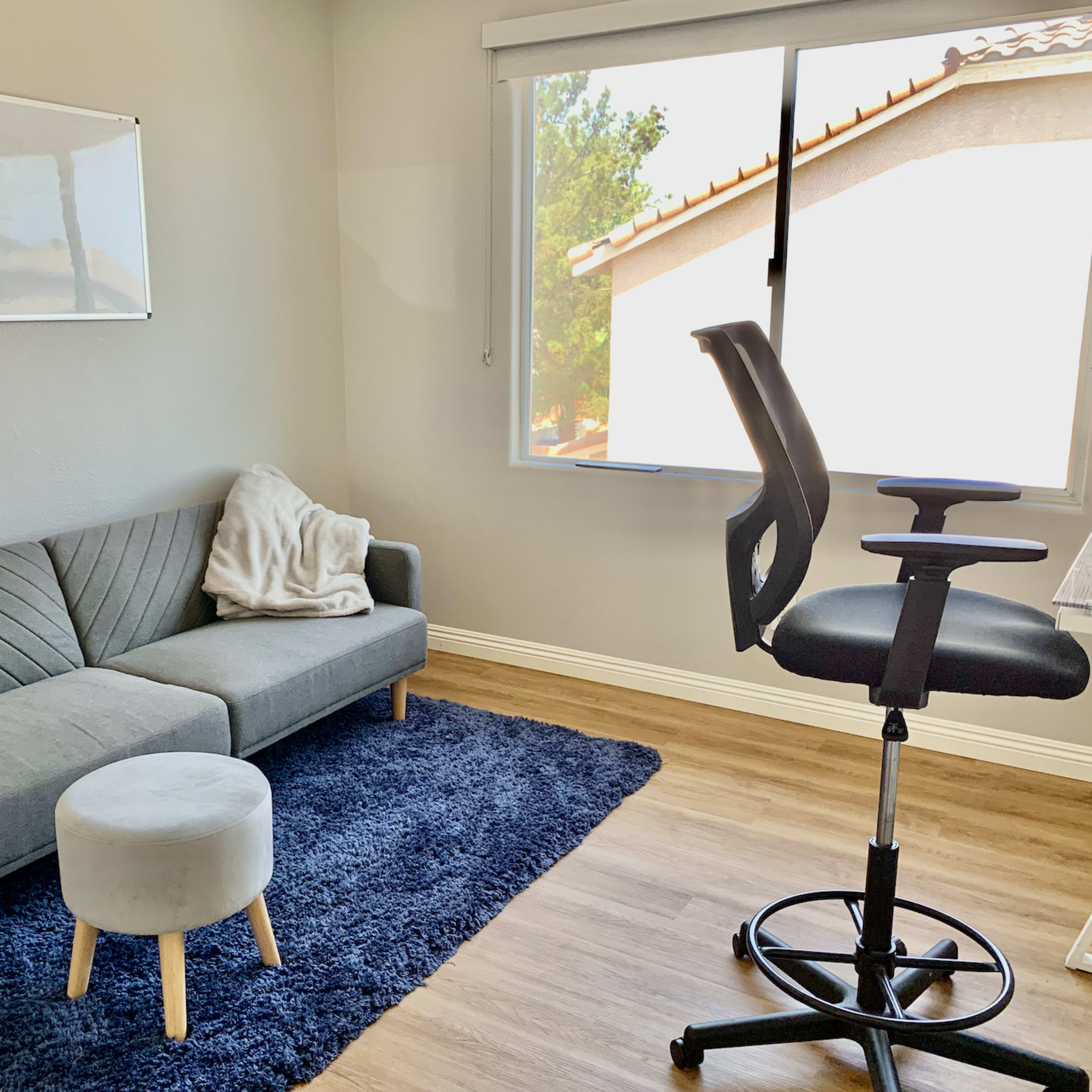 A minimalist office space featuring a gray sofa with a light blanket, a round stool, a swivel chair, and a desk by a window.