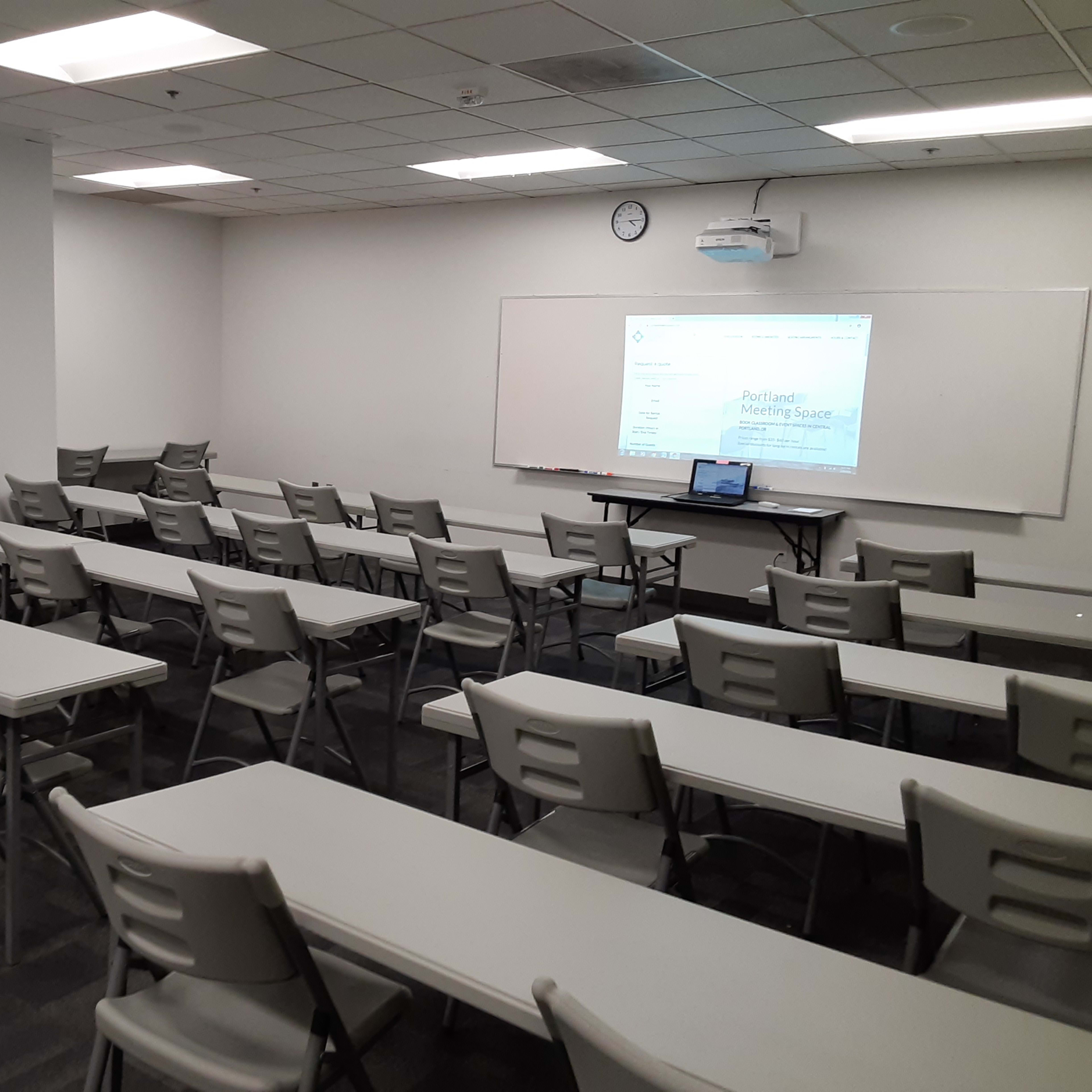 A classroom with multiple rows of folding tables and chairs, a projector setup, and a whiteboard at the front.