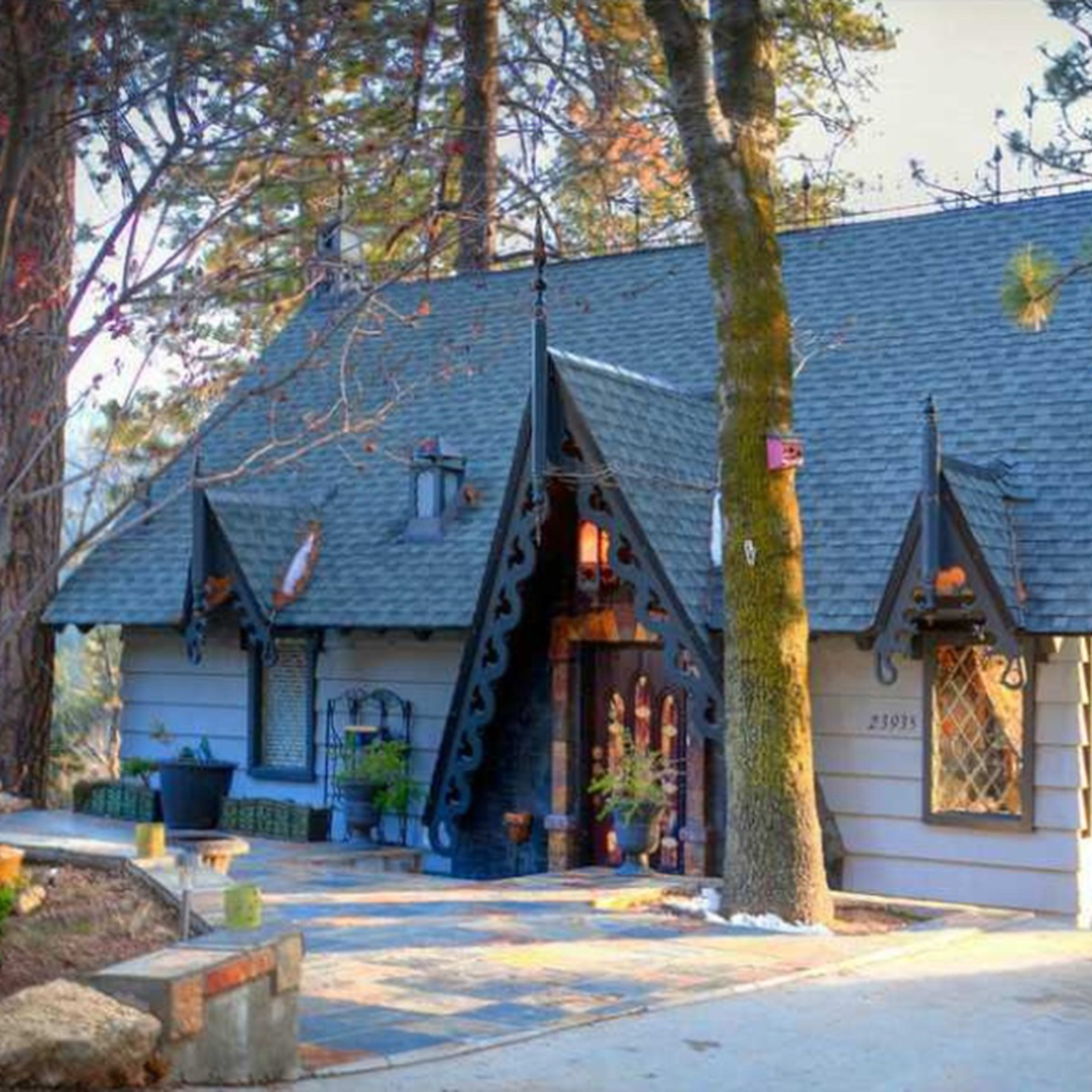 A small, intricately designed house with a steep roof and decorative elements, surrounded by tall trees and a stone pathway.