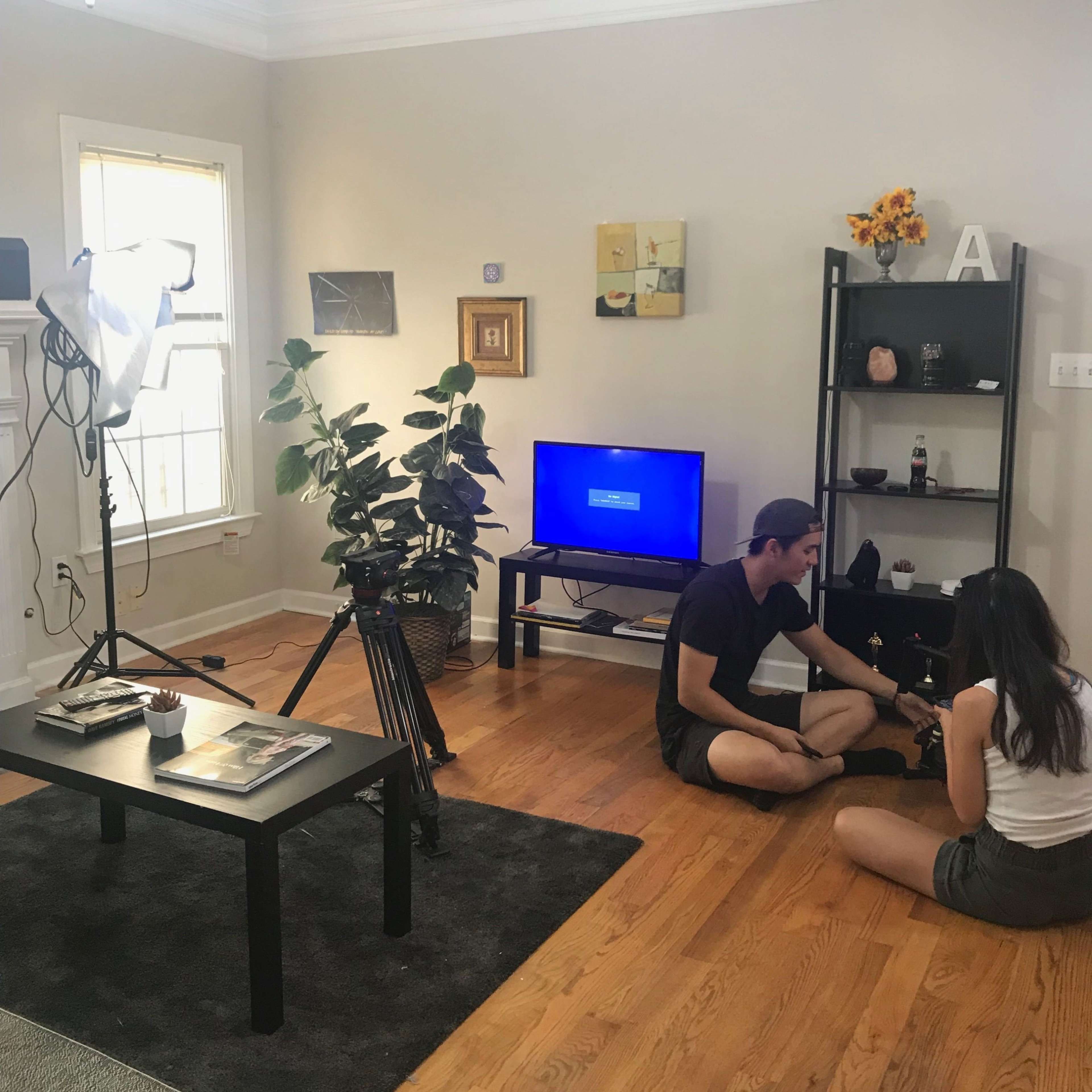 Two people are setting up equipment in a well-lit living room with a television and a bookshelf.