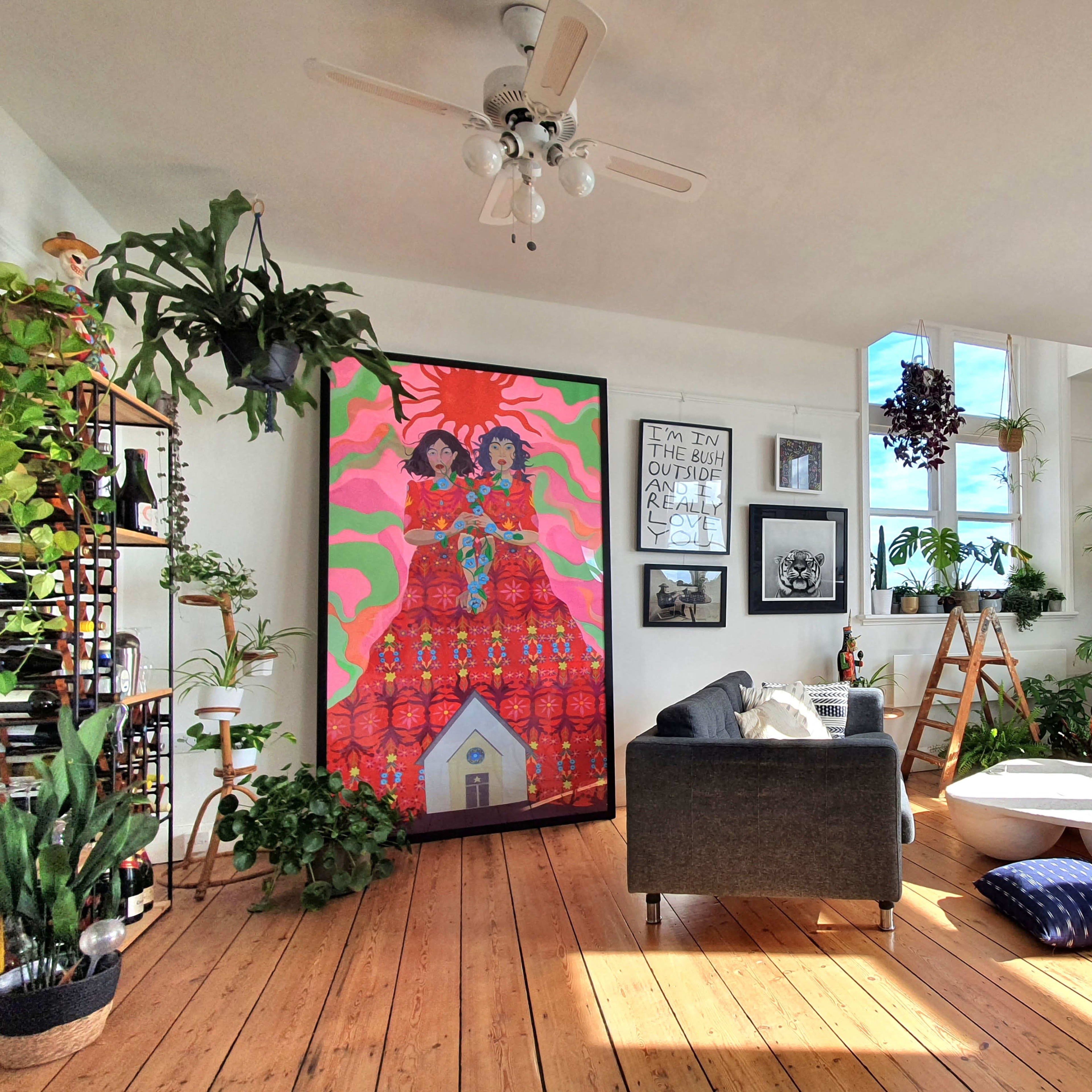 A bright, modern living room with large windows, featuring a colorful mural, houseplants, and a mix of furniture including a sofa and a coffee table.