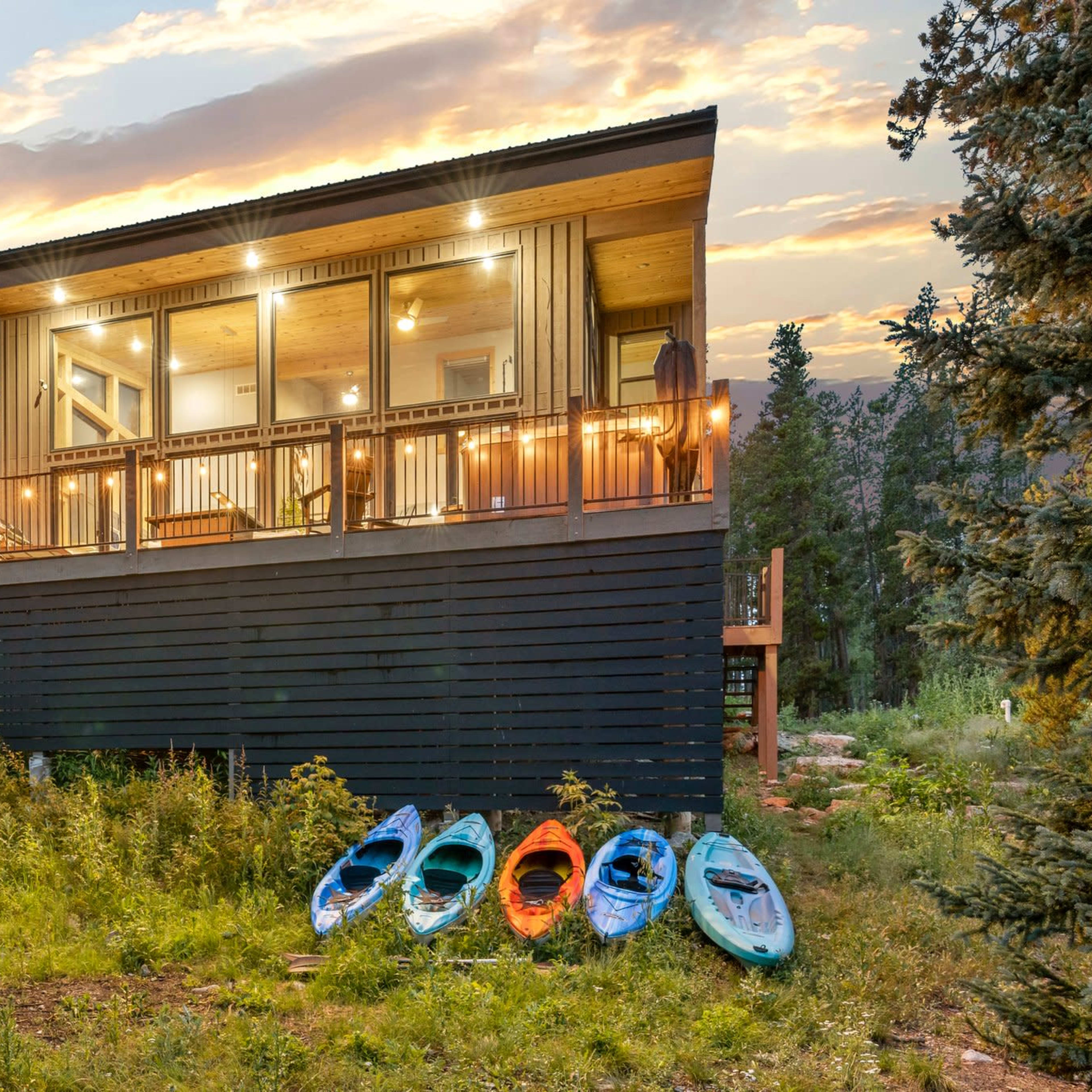 A modern cabin with large windows is situated on a grassy area, featuring a deck and several kayaks lined up nearby.