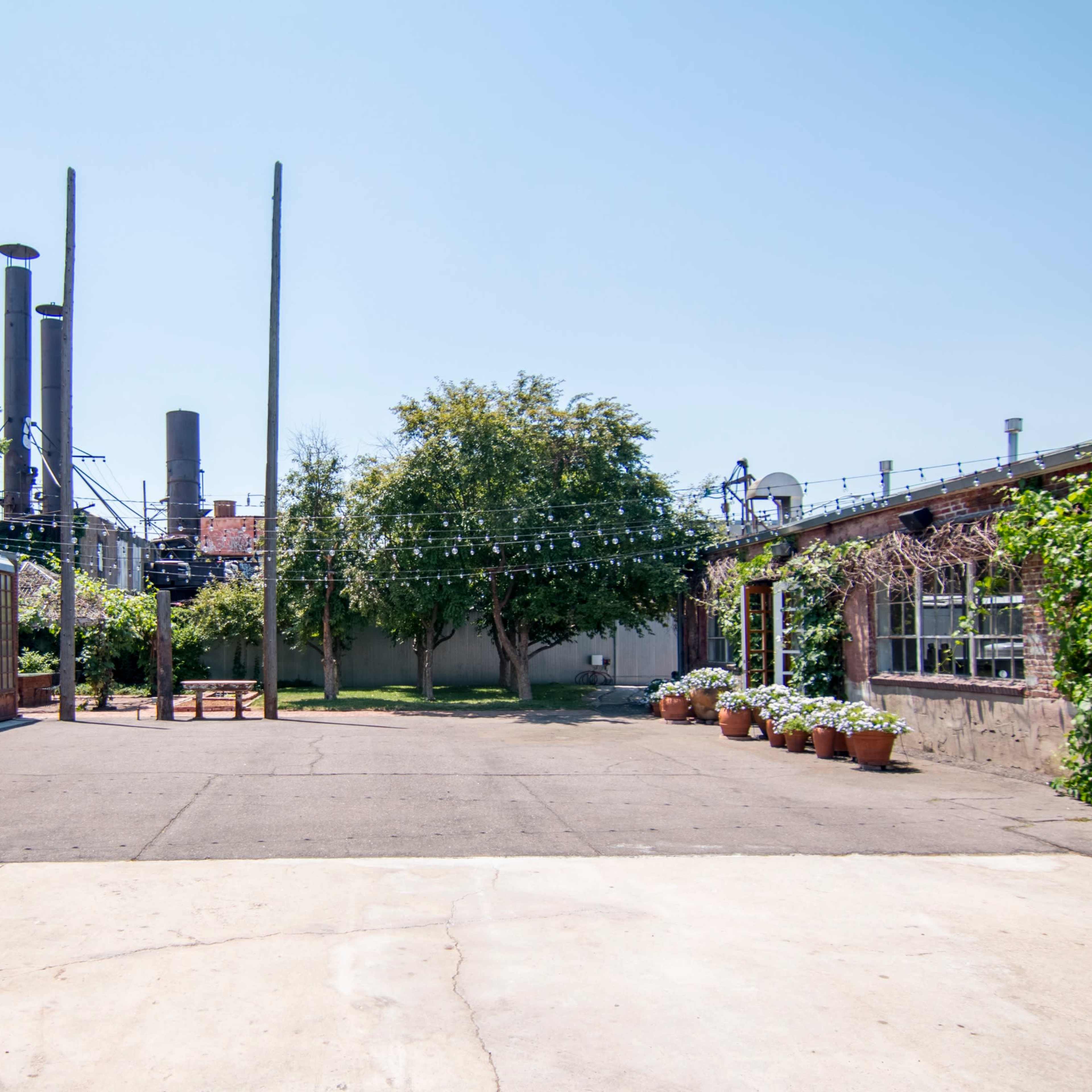 An open courtyard bordered by buildings, with potted plants and utility poles in the background under a clear sky.