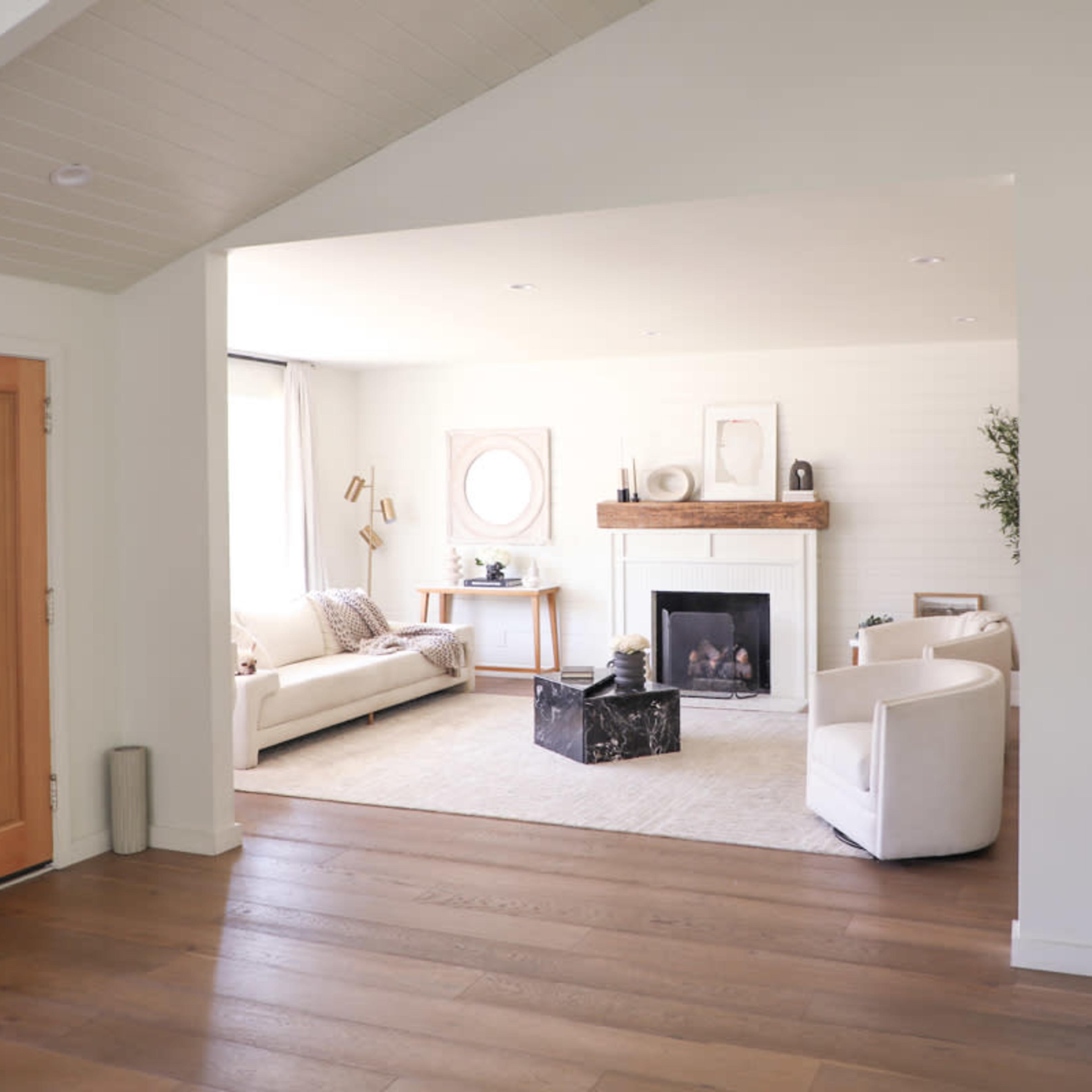 An open living room with a fireplace, two white sofas, a coffee table, and large windows allowing natural light to enter.
