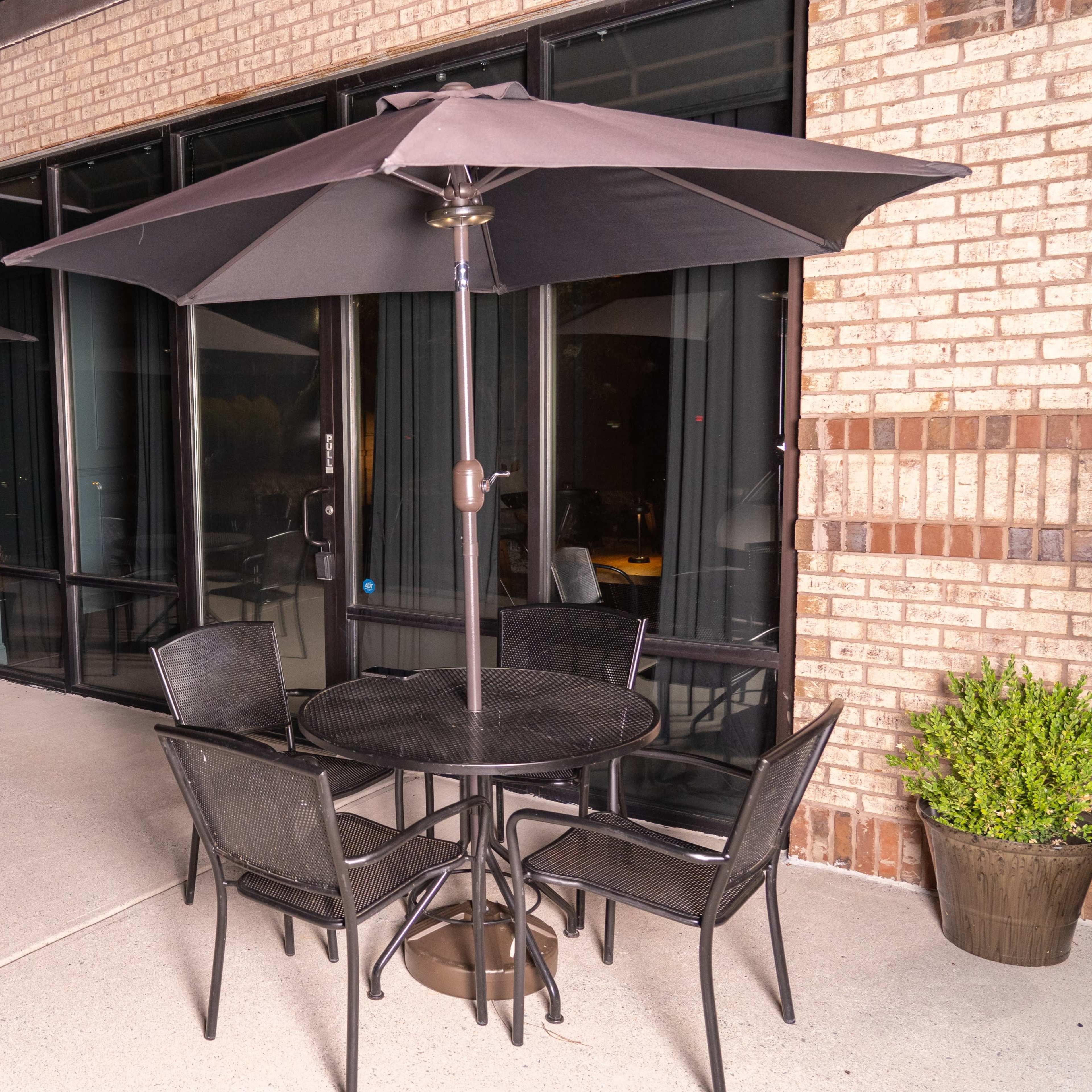 An outdoor seating area with black metal tables and chairs under large umbrellas, situated next to a brick building.