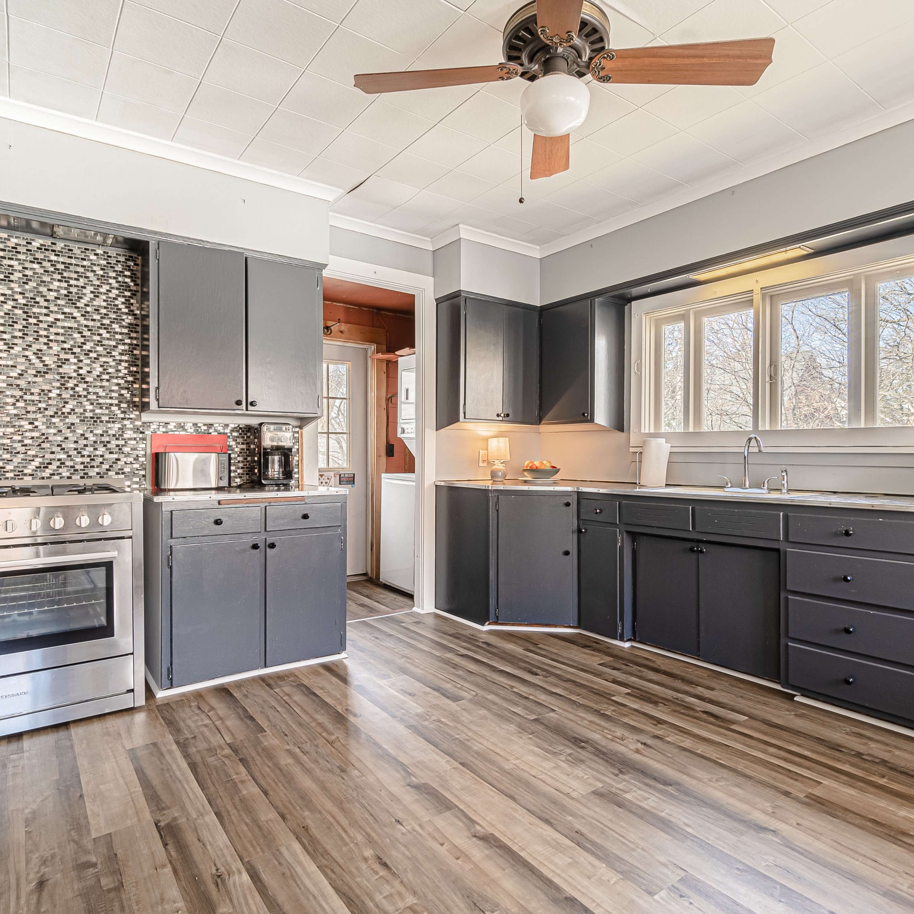 The kitchen features dark cabinets, a stainless steel stove, and a mosaic tile backsplash, with natural light streaming through the window.