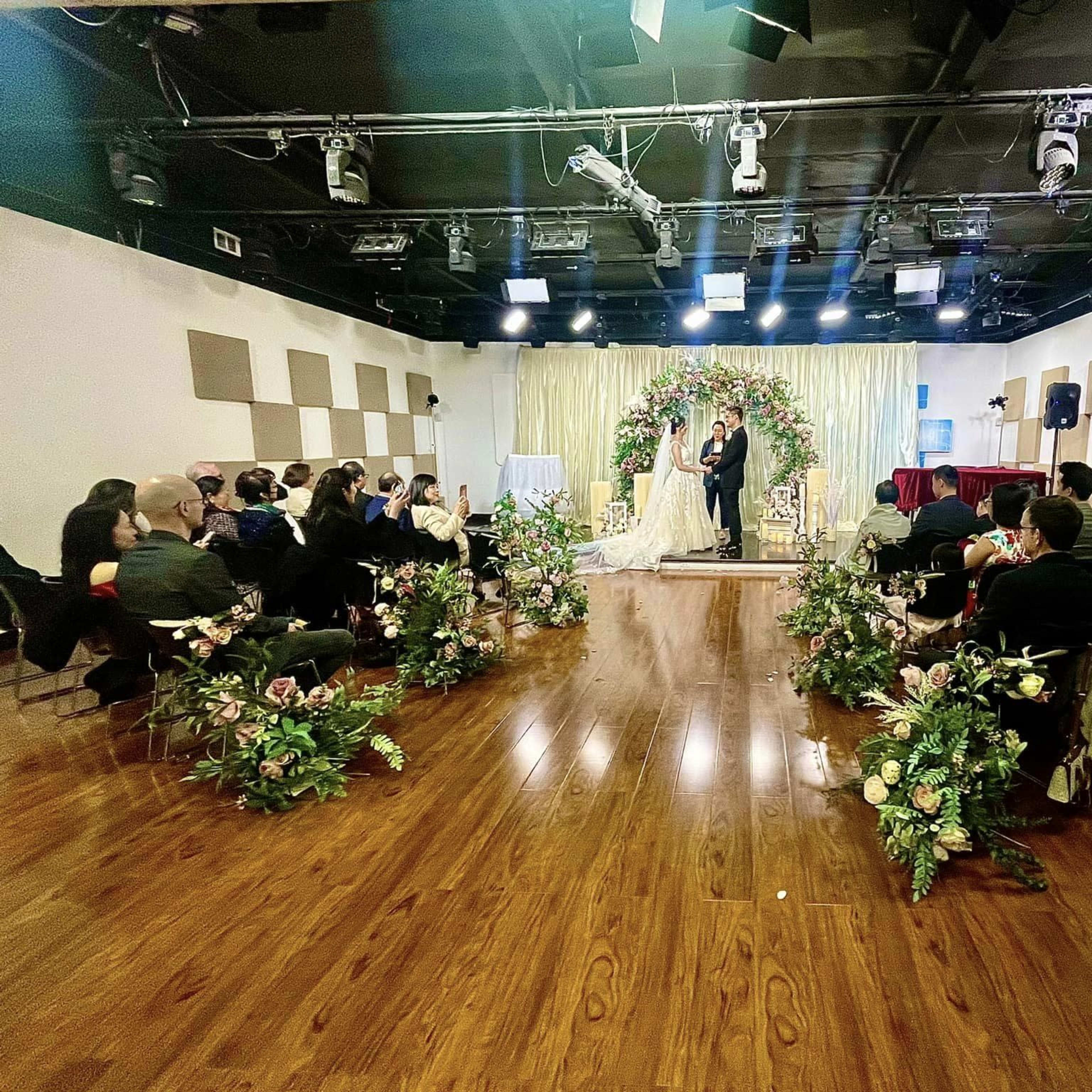A couple stands in front of a floral arch while guests watch from arranged seating in a decorated event space.