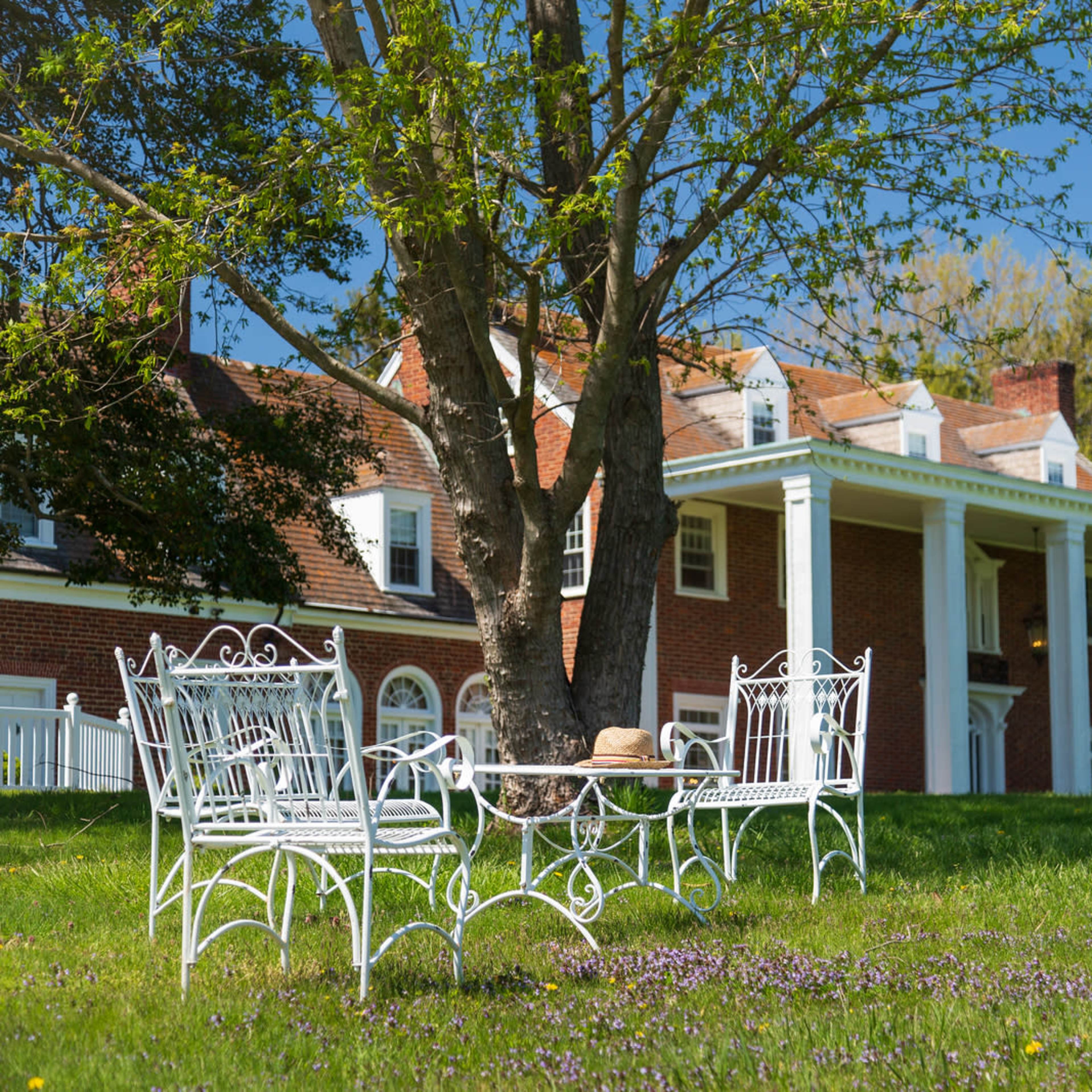A white wrought iron table and chairs are placed under a tree in front of a large brick house with white columns.
