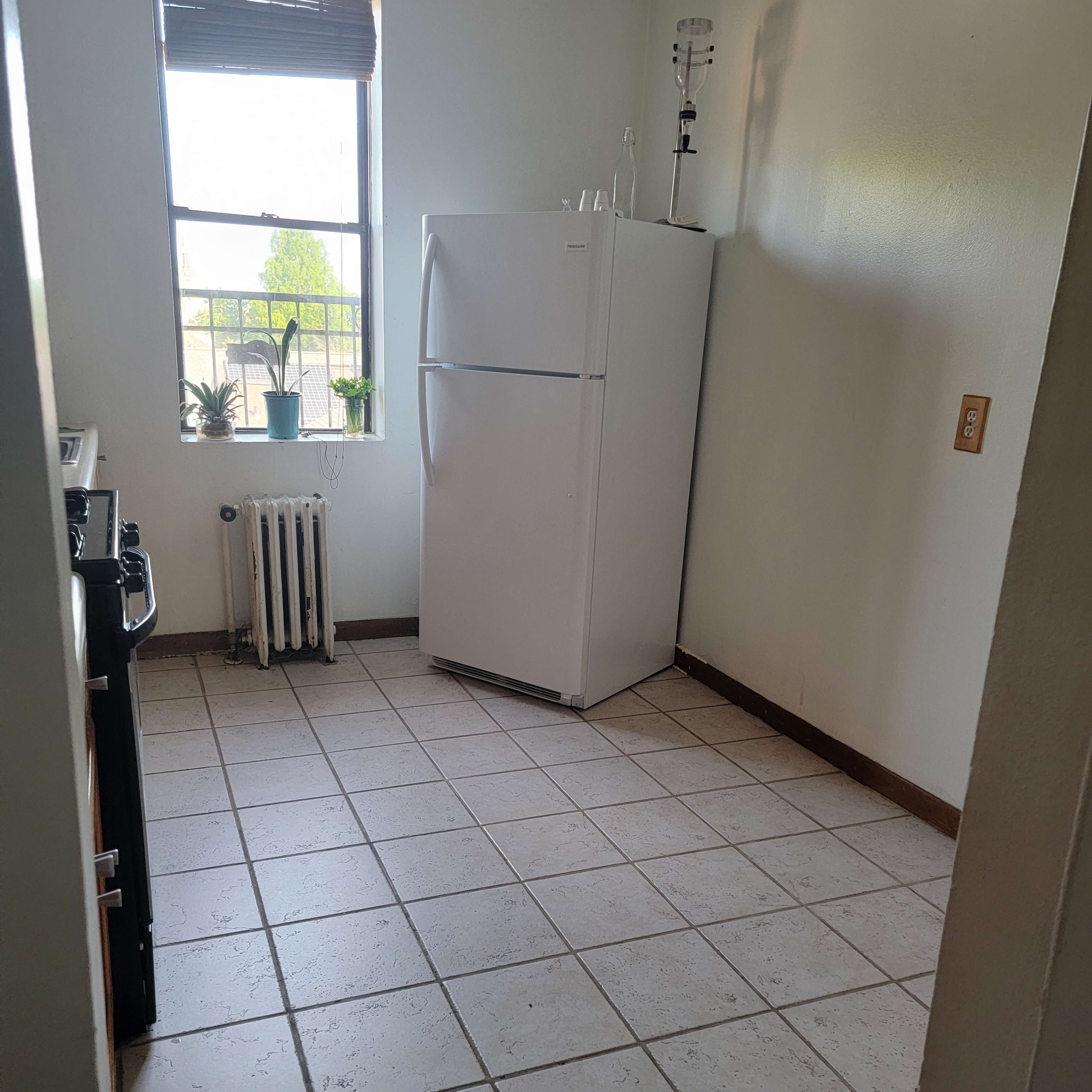 A small kitchen features a white refrigerator, a stove, and a window with plants on the sill.