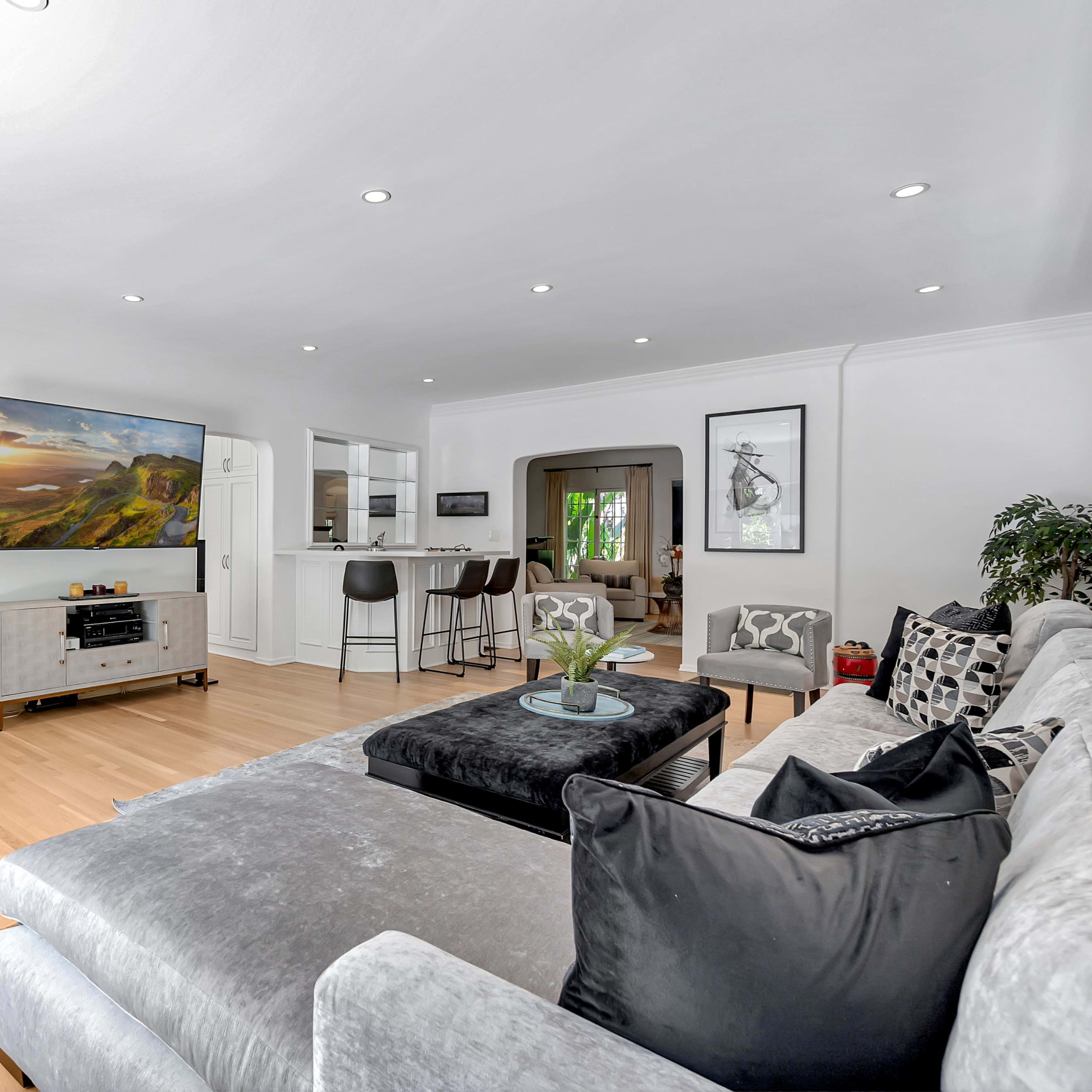 A modern living room features a sectional sofa, a coffee table, and a large television, with an open kitchen visible in the background.
