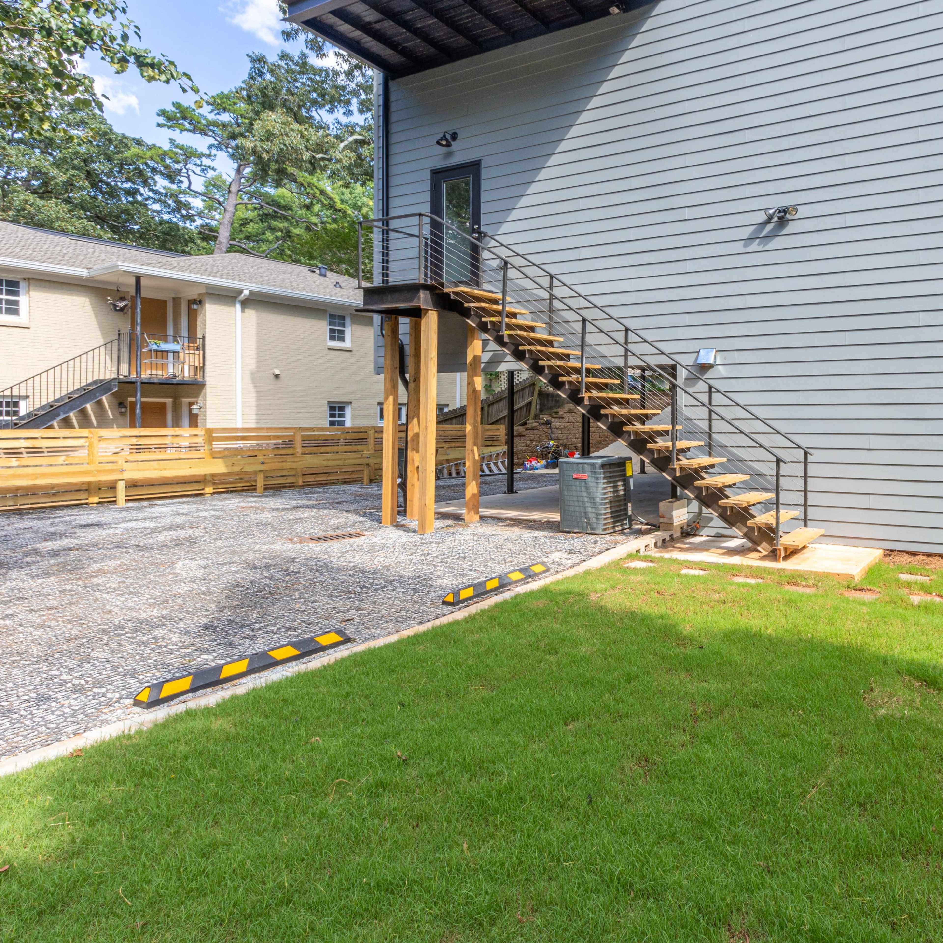 The exterior of a modern gray house with wooden stairs leading to a second floor, situated next to a gravel parking area and a landscaped yard.