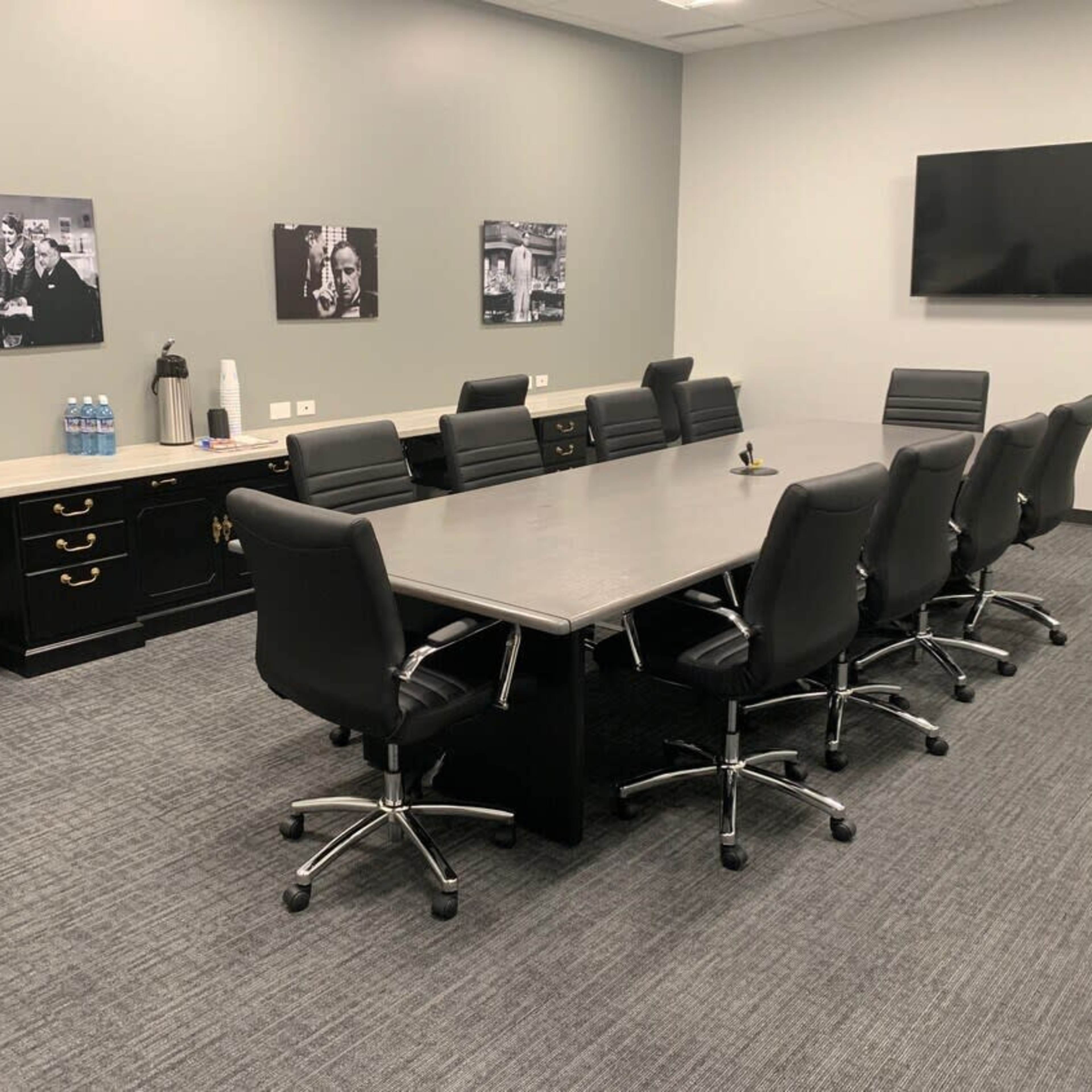 A modern conference room with a long table surrounded by black swivel chairs, a large screen on the wall, and water bottles on a counter in the background.